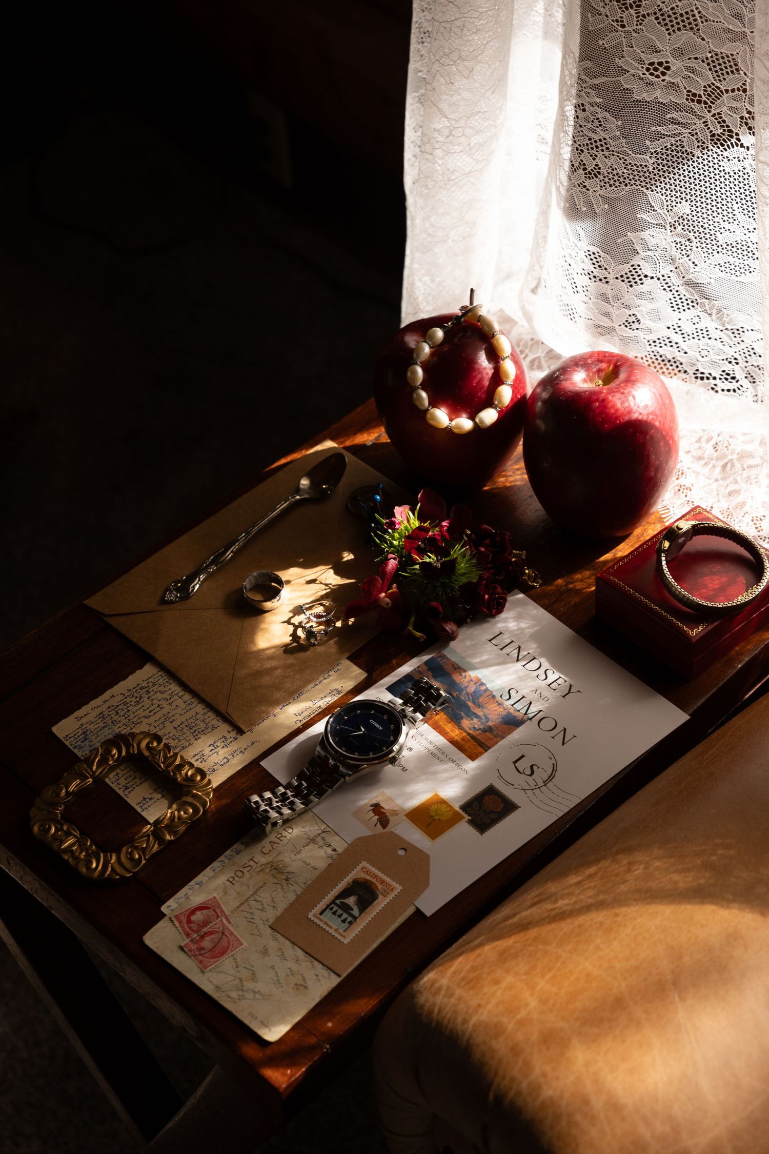 Vintage wooden table with apples, a watch, jewelry, flowers, and papers in warm sunlight near lace curtains.