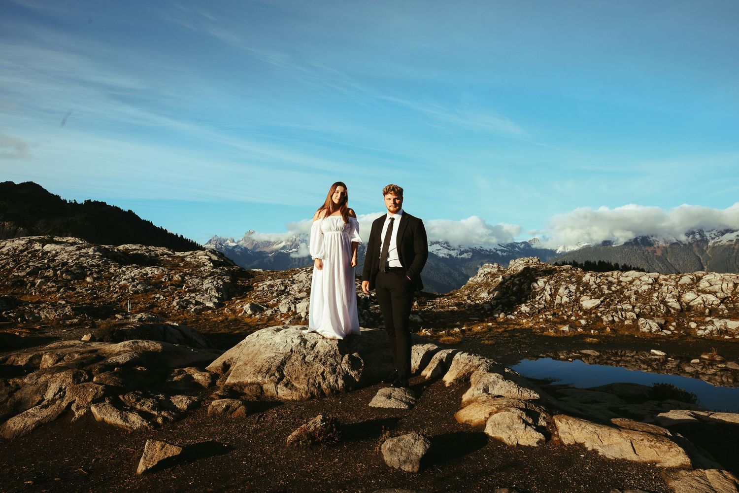 Bride in a white gown and groom in a dark suit stand hand in hand on a rocky mountain ridge with distant peaks and blue sky.