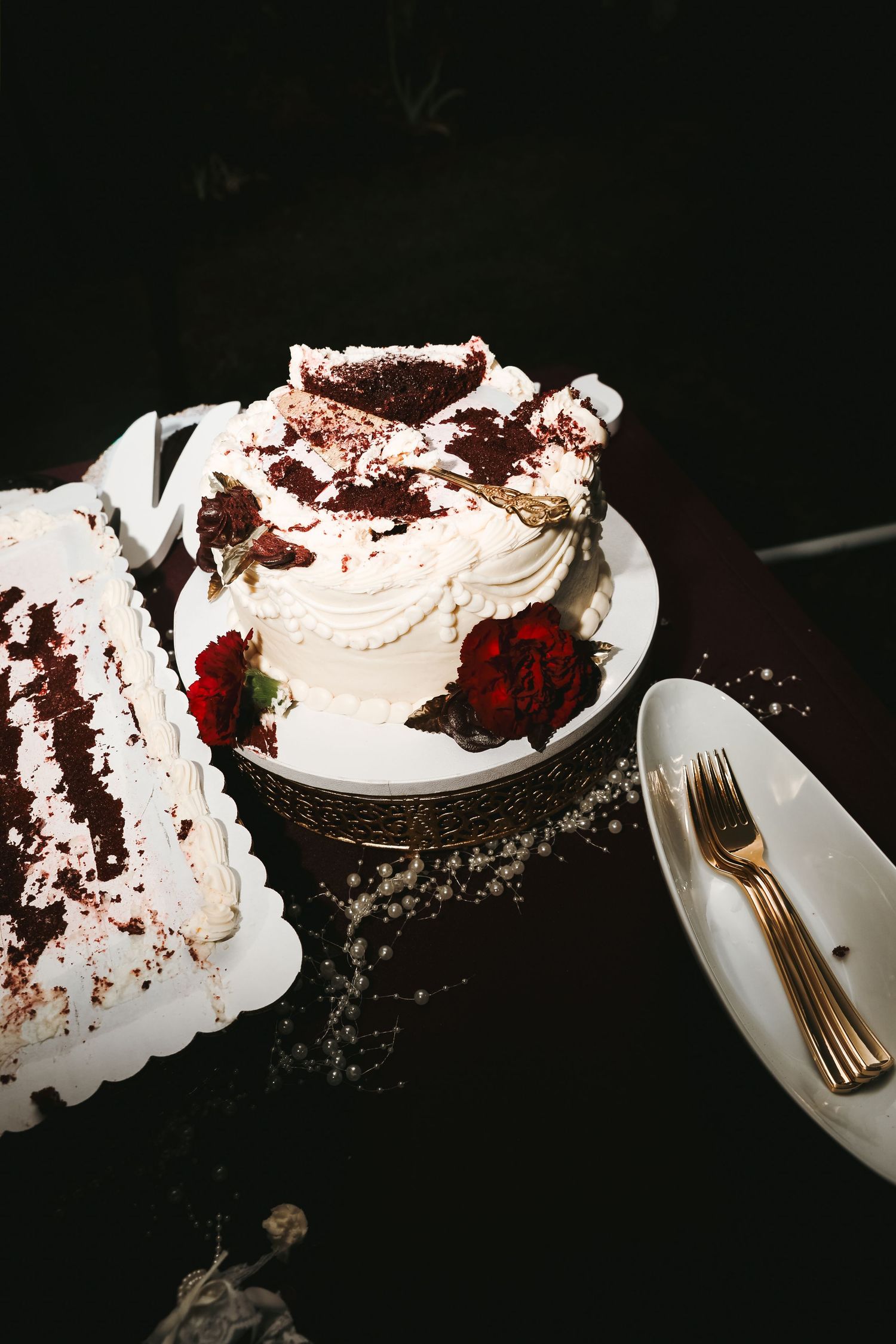 White frosted wedding cake with red velvet crumbs and dark red roses on a table with gold forks and a sliced sheet cake.