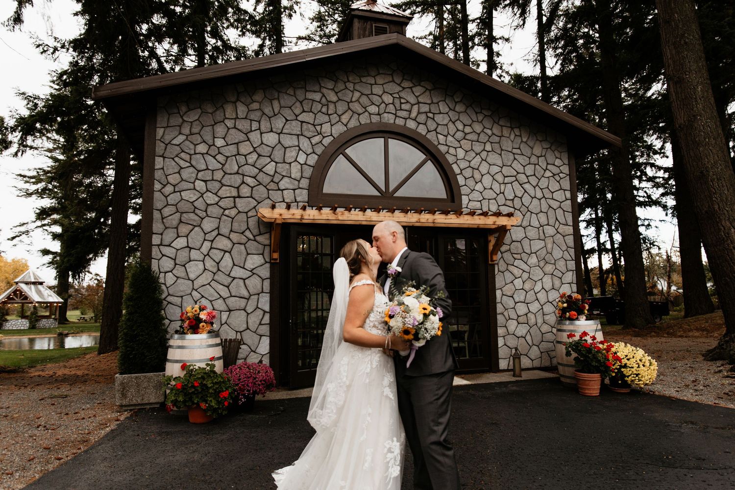 Bride and groom share a romantic kiss in front of a charming stone chapel surrounded by fall foliage.