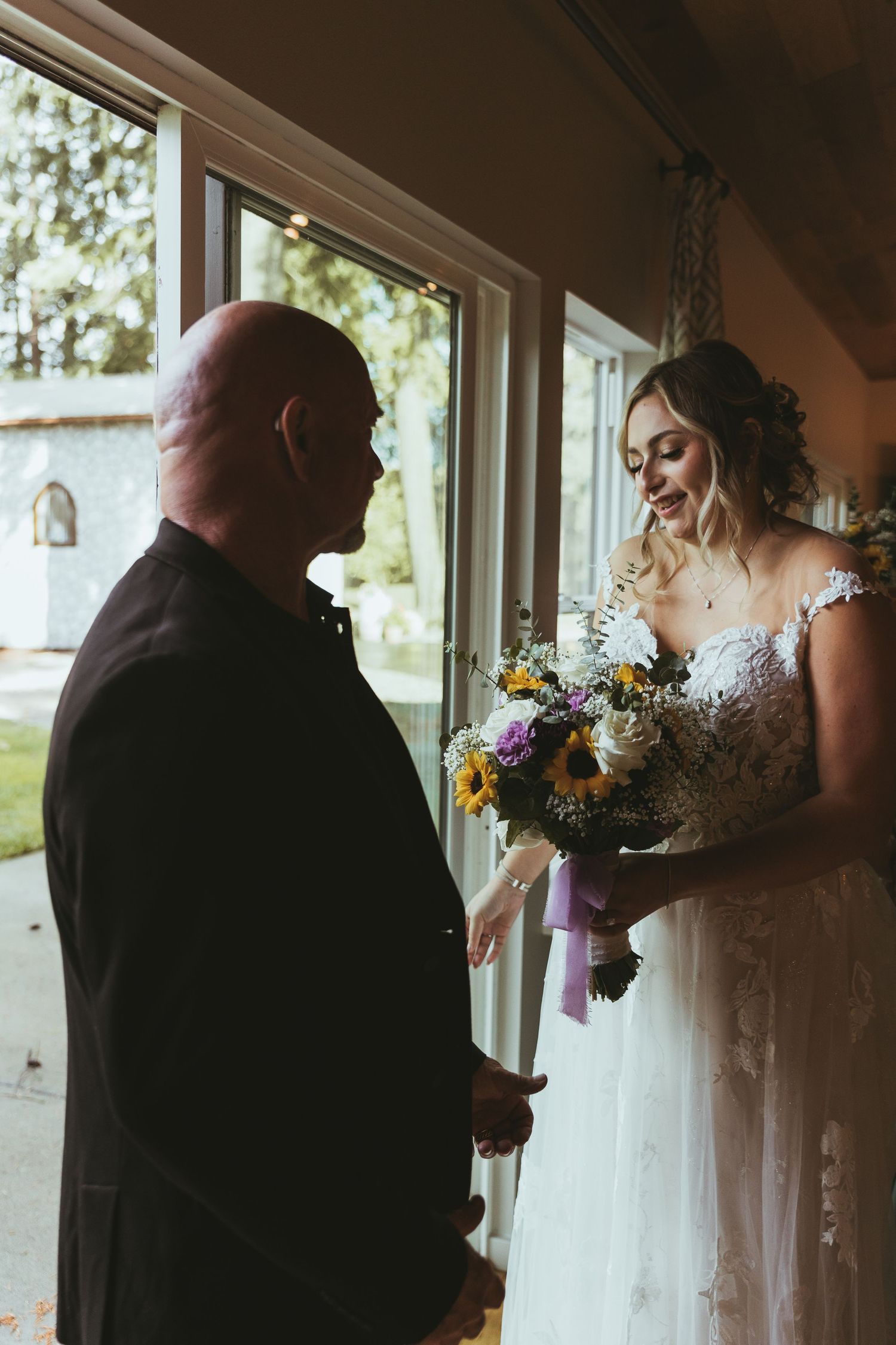 Bride in lace wedding gown smiles while holding a colorful bouquet during an intimate first look moment.