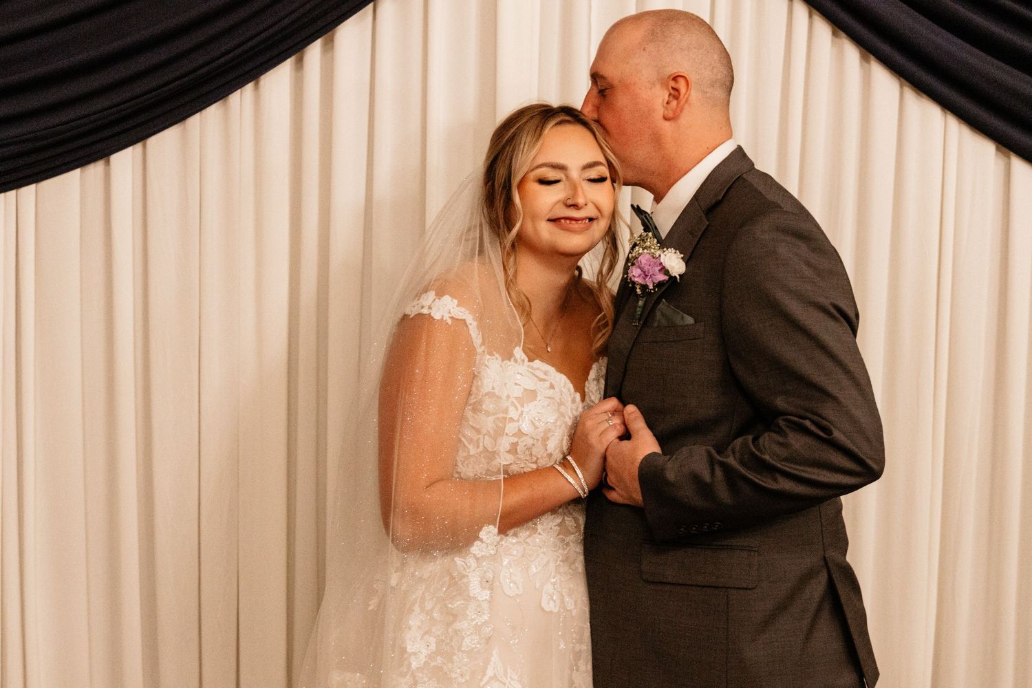 Groom kissing smiling bride on the cheek while holding hands in front of elegant white drape backdrop.