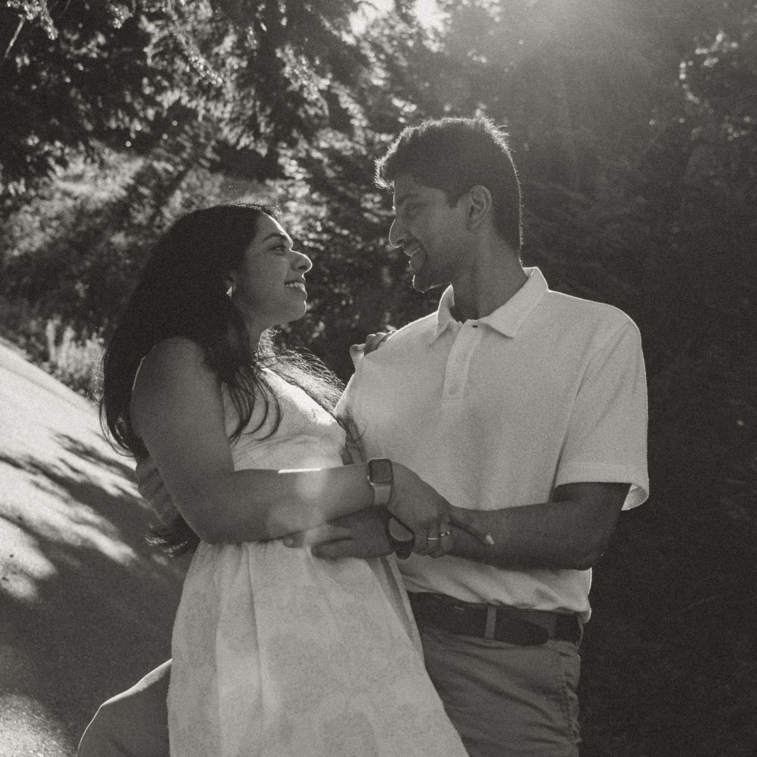 Black and white photo of a couple gazing at each other outdoors, bathed in warm sunlight with trees in the background.