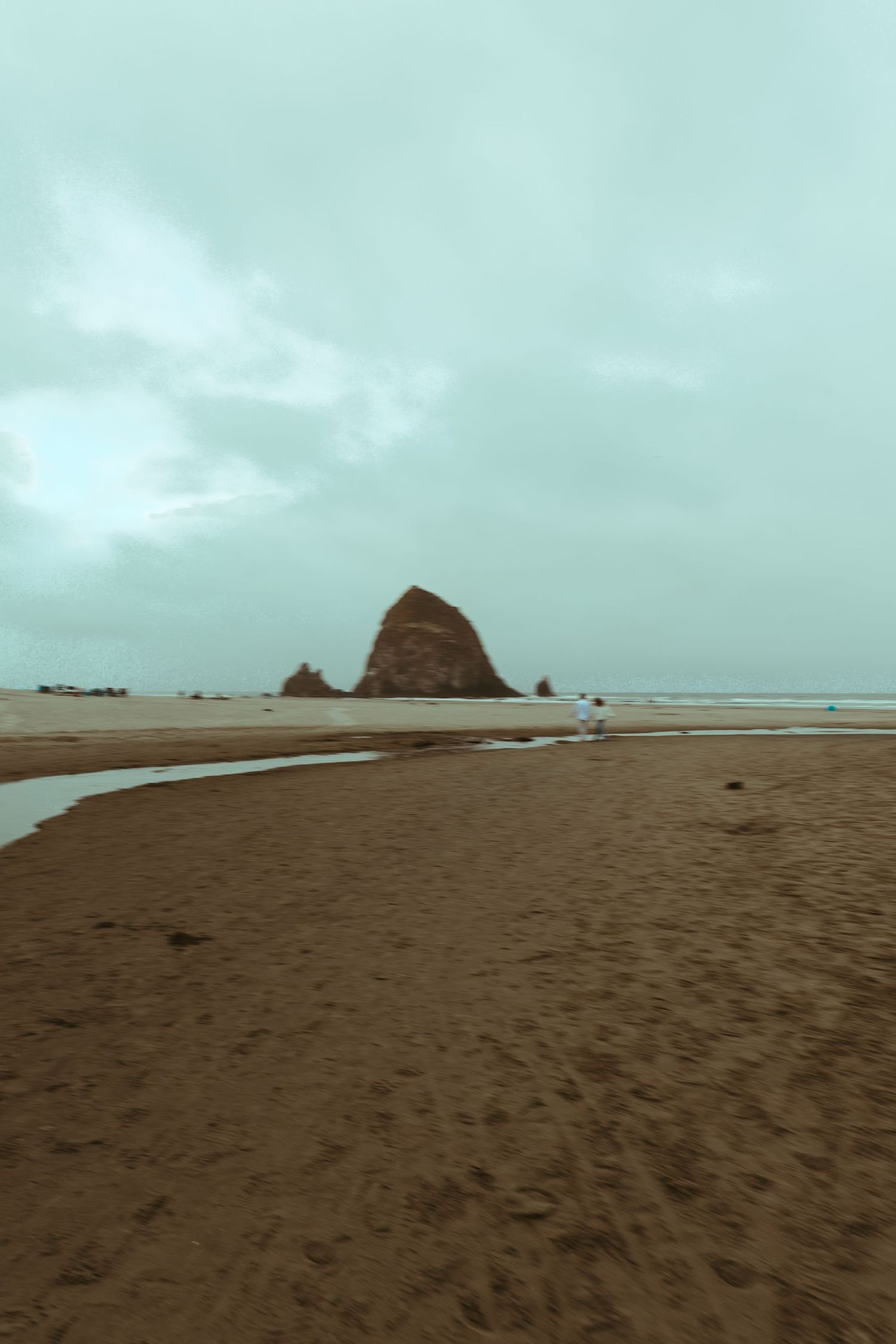 Overcast sky over a wet sandy beach with a tidal stream leading toward a large sea stack rock rising from the shoreline in the distance.