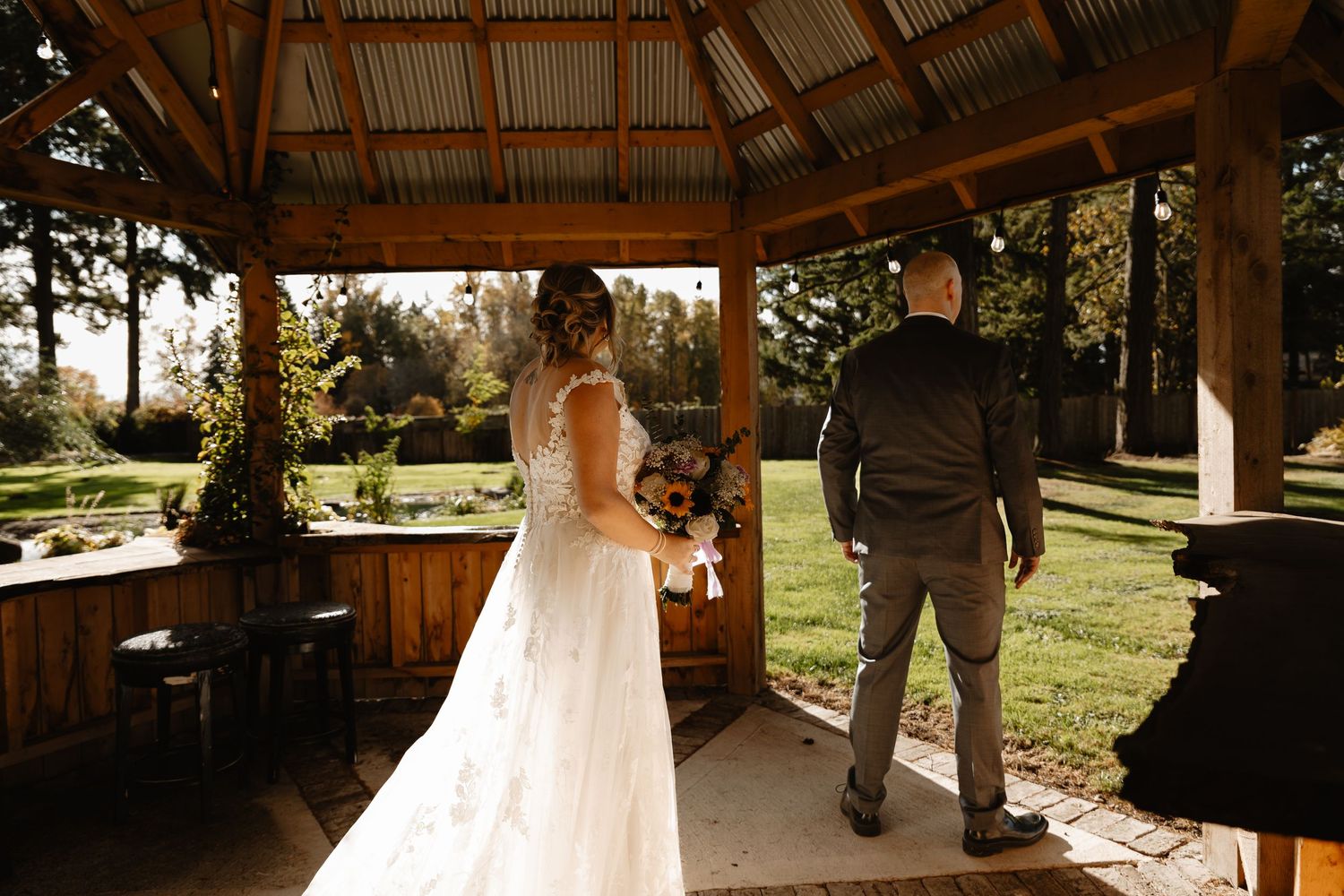 Bride holding floral bouquet stands with groom inside wooden gazebo during wedding first look reveal.