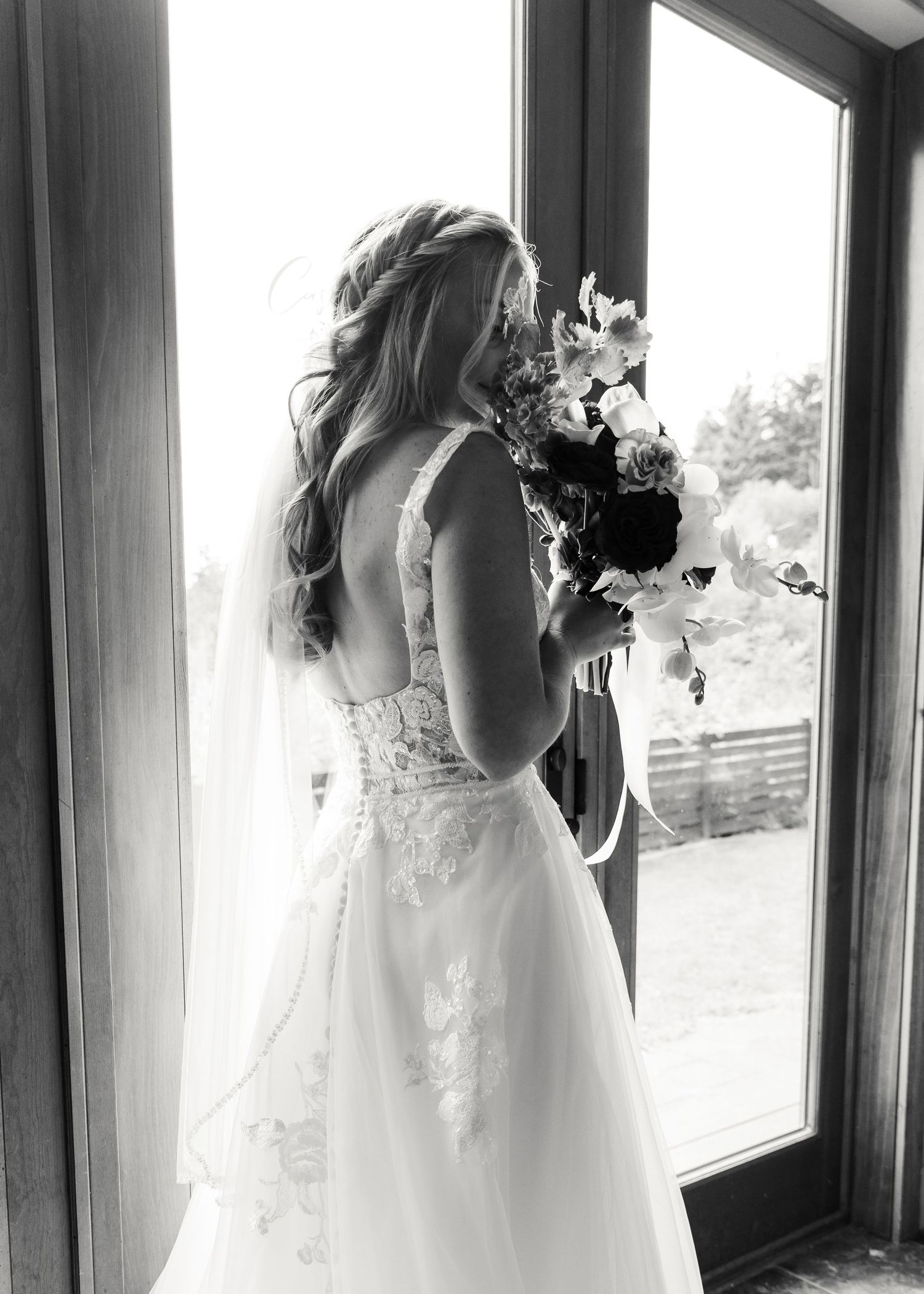 Bride in lace wedding gown holding bouquet, standing by window in black and white photo.