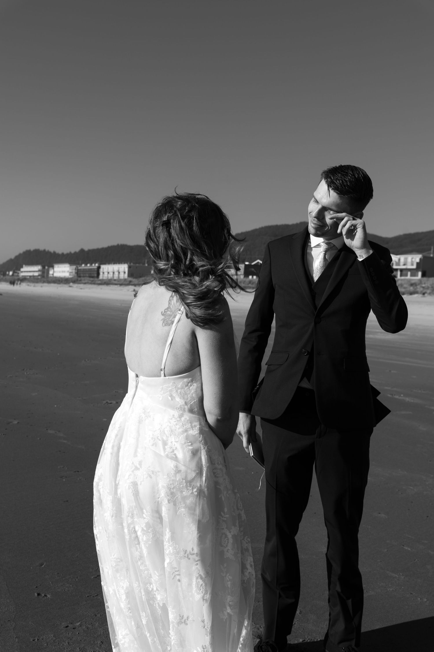 Black and white photo of a bride and groom holding hands on a beach, groom in suit, bride in backless lace gown.