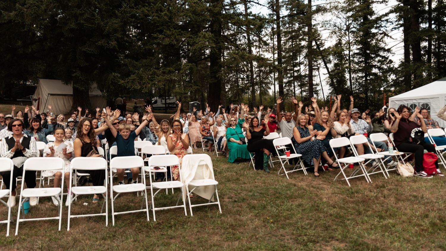 Outdoor wedding reception guests cheering on white folding chairs on green lawn surrounded by trees.