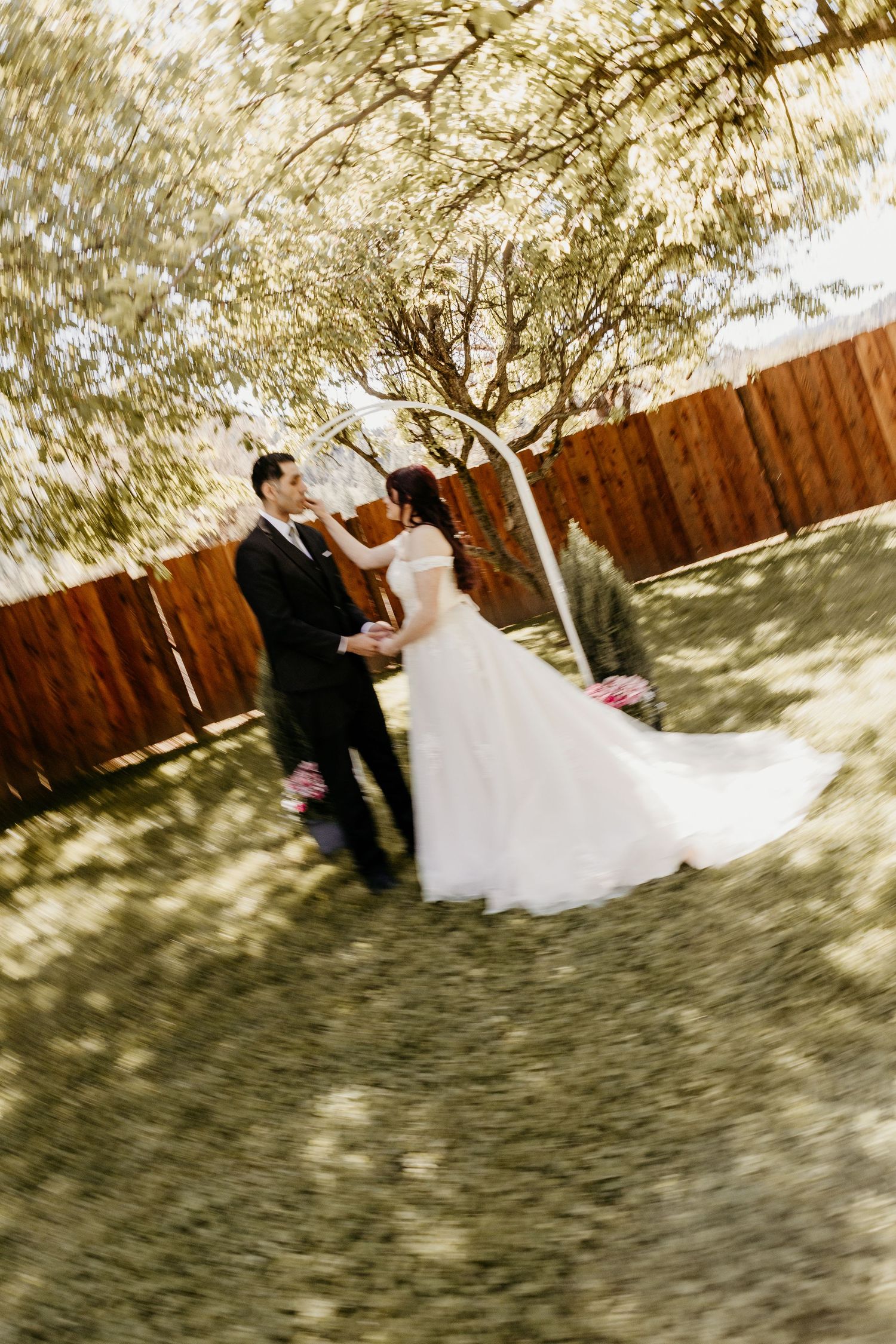 Bride and groom share a romantic moment outdoors, surrounded by lush green trees and a wooden fence backdrop.