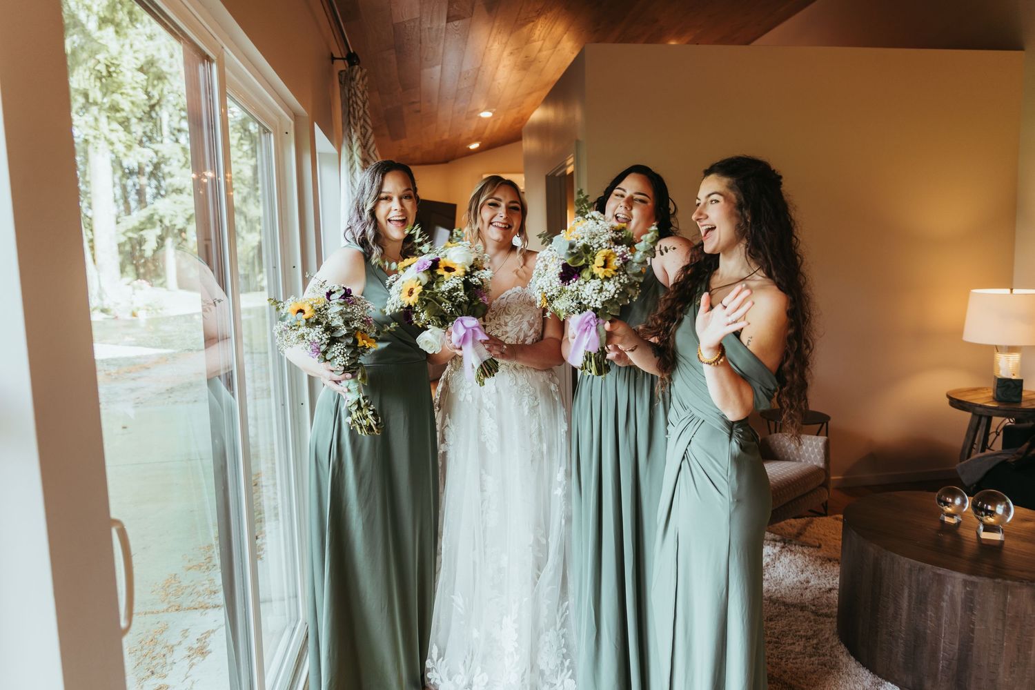 Bride and three bridesmaids in sage green dresses laugh together while holding vibrant floral bouquets by a window.