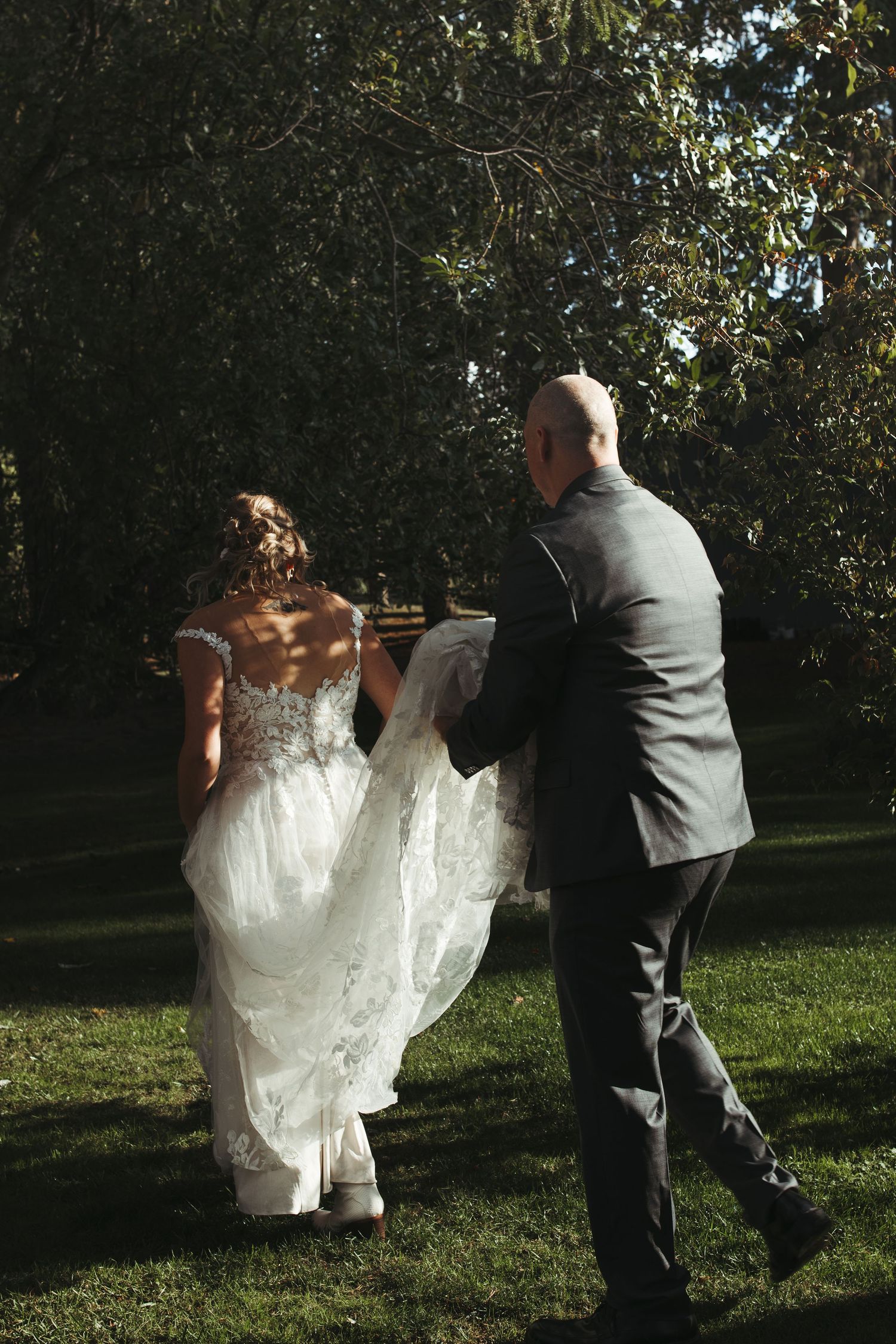 Bride and groom walking hand in hand through a sunlit garden, groom carrying the brides lace train.