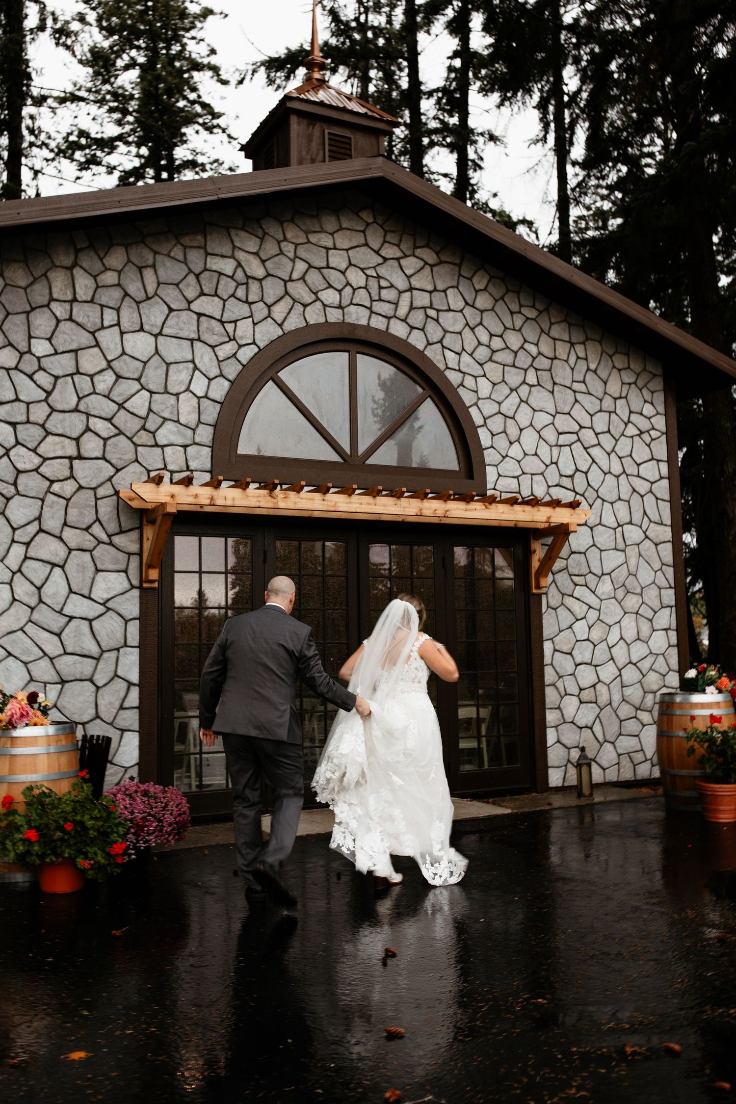 Bride and groom walking hand in hand toward stone chapel entrance on a rainy wedding day.