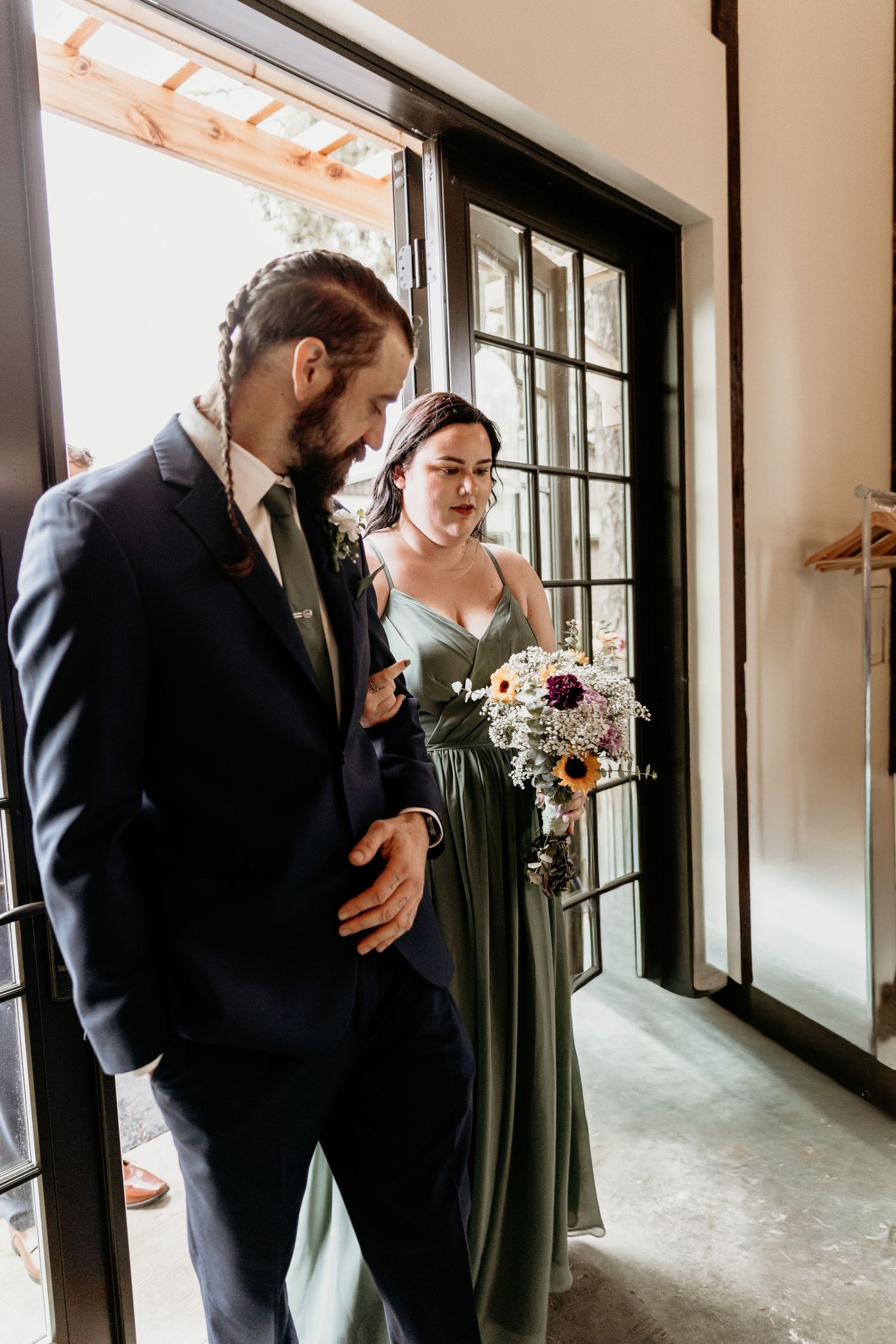A groomsman in a navy suit escorts a bridesmaid in a sage green gown holding a wildflower bouquet.