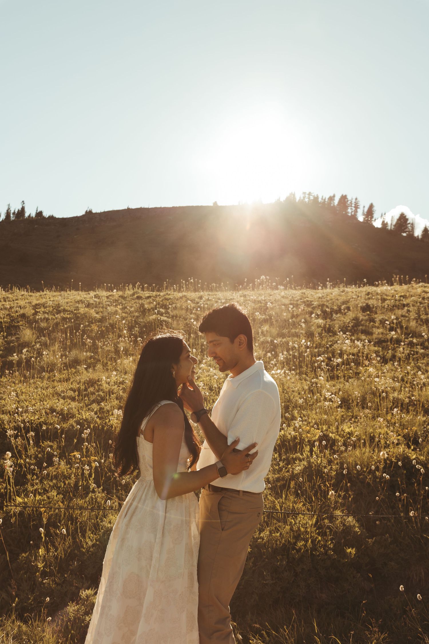 Couple embracing in a sunlit meadow at golden hour, with warm backlight and rolling hills in the background.