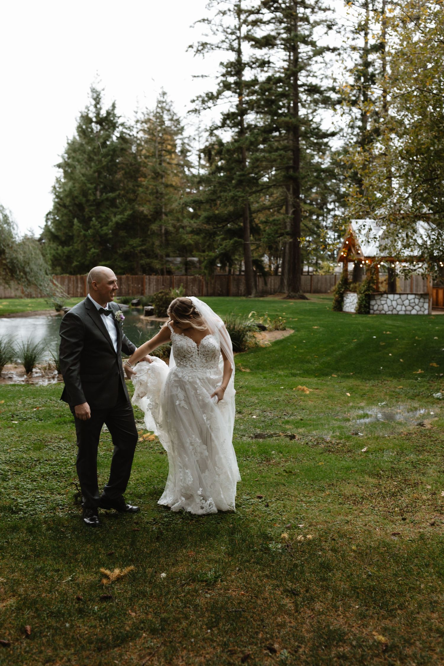 Groom helps bride with veil while walking on lush green lawn.