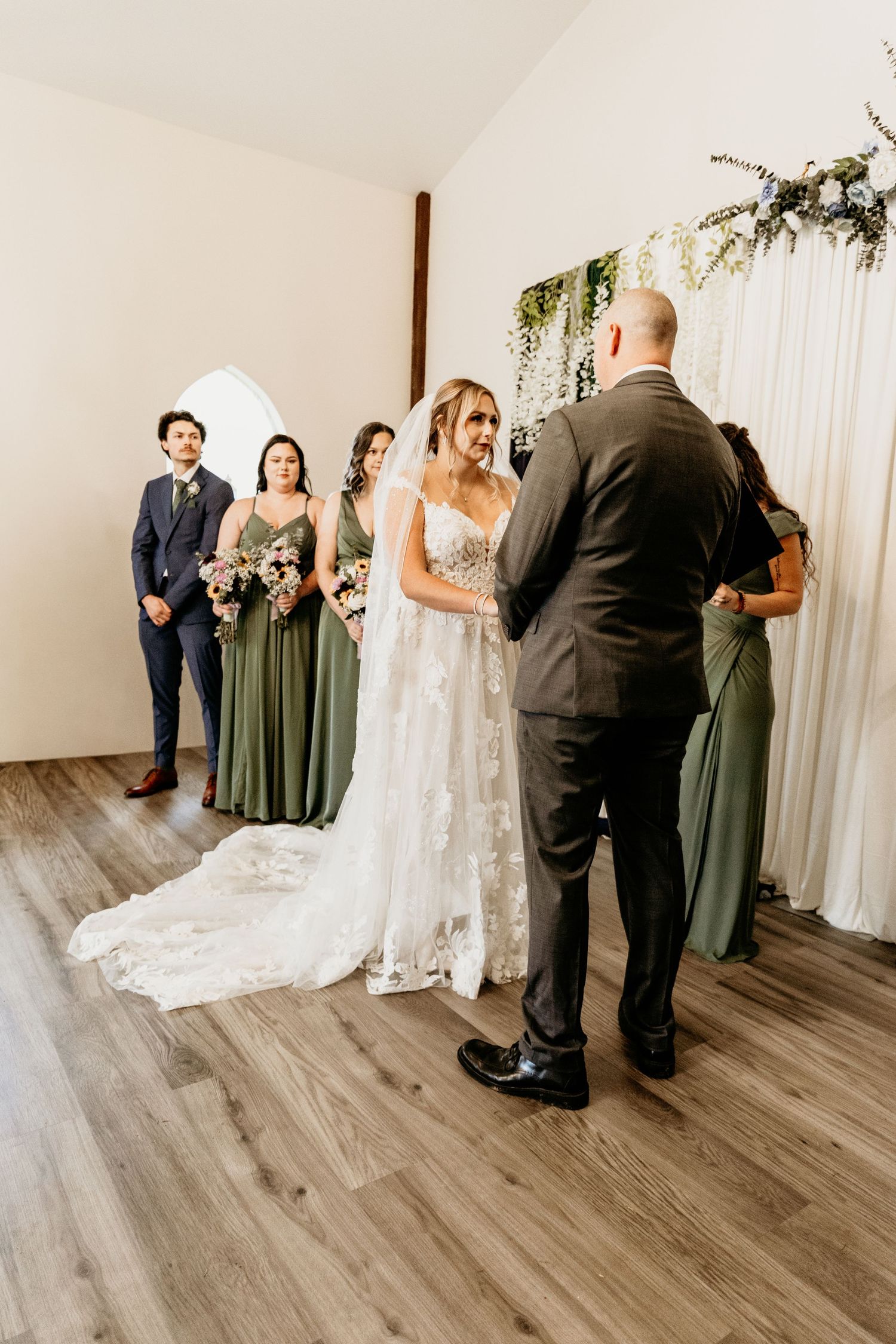 Bride in lace gown exchanges vows with groom at indoor wedding ceremony with bridesmaids in sage green dresses.