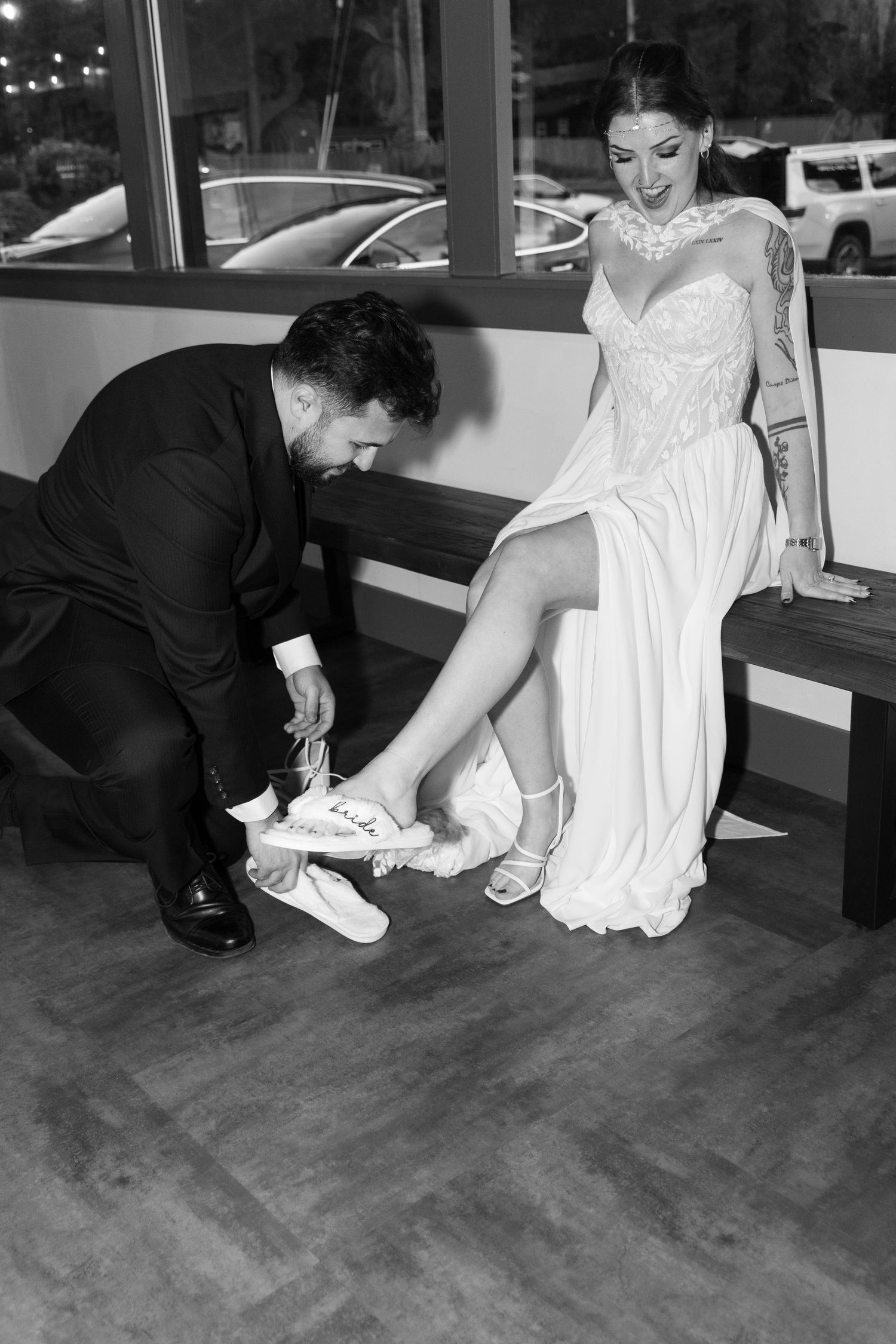Black and white photo of a groom in a suit kneeling to help a bride in a lace wedding gown change into sneakers while seated on a bench.
