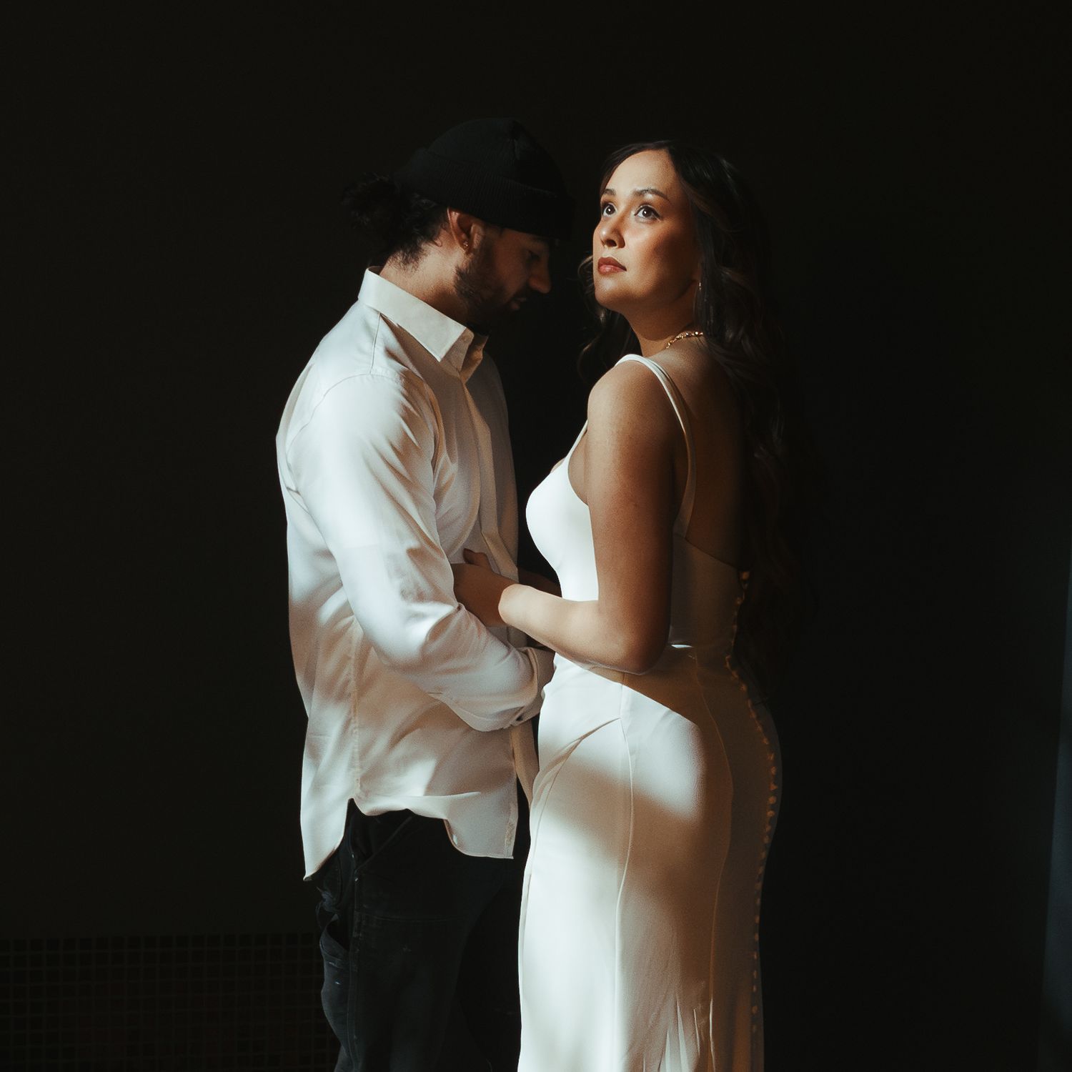 A couple in elegant white attire embraces intimately against a dark background in a moody romantic portrait.