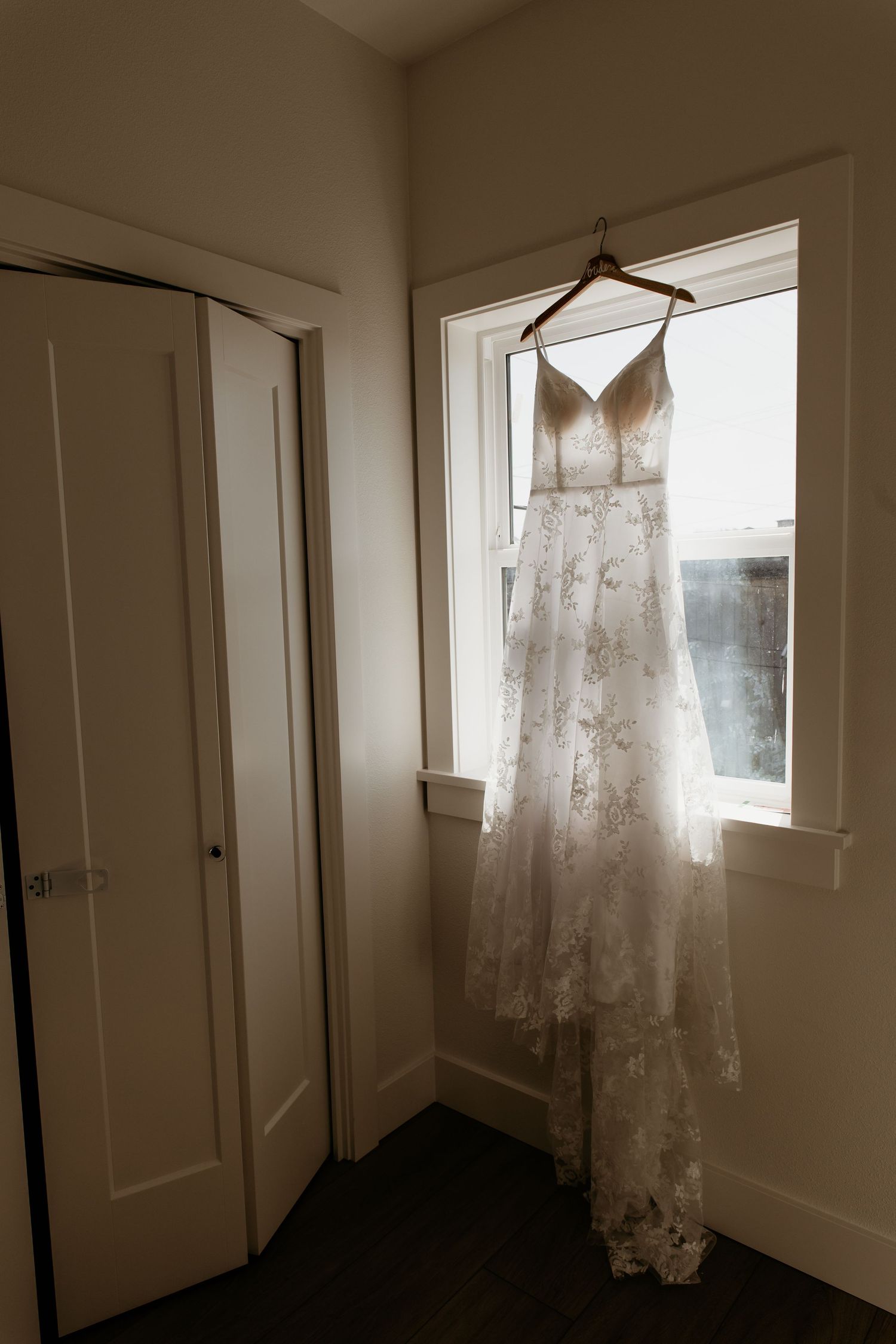 Elegant lace wedding dress hanging on a wooden hanger in front of a bright window in a bridal suite.