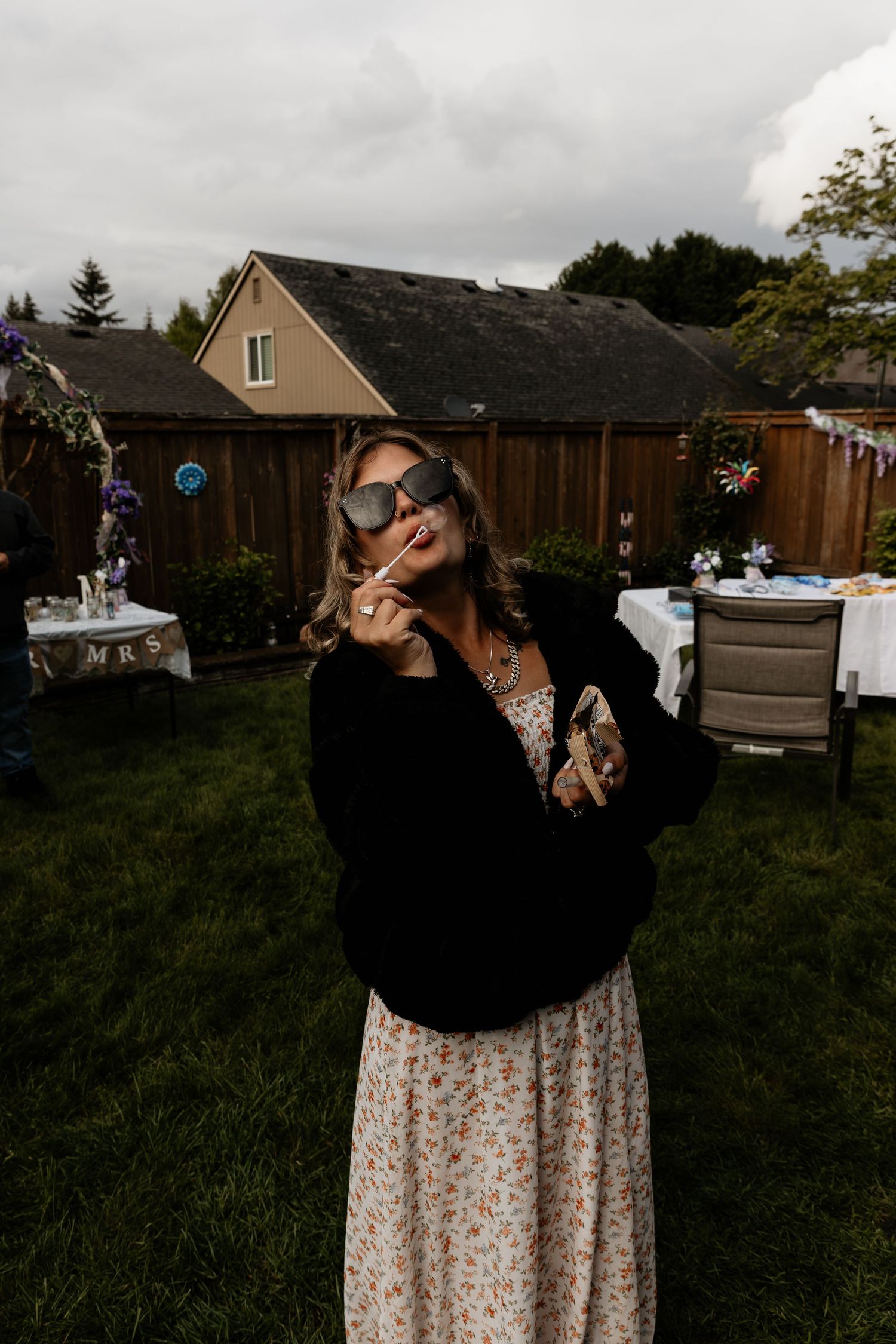 Woman in floral dress and black cardigan poses with oversized sunglasses at an outdoor backyard party.