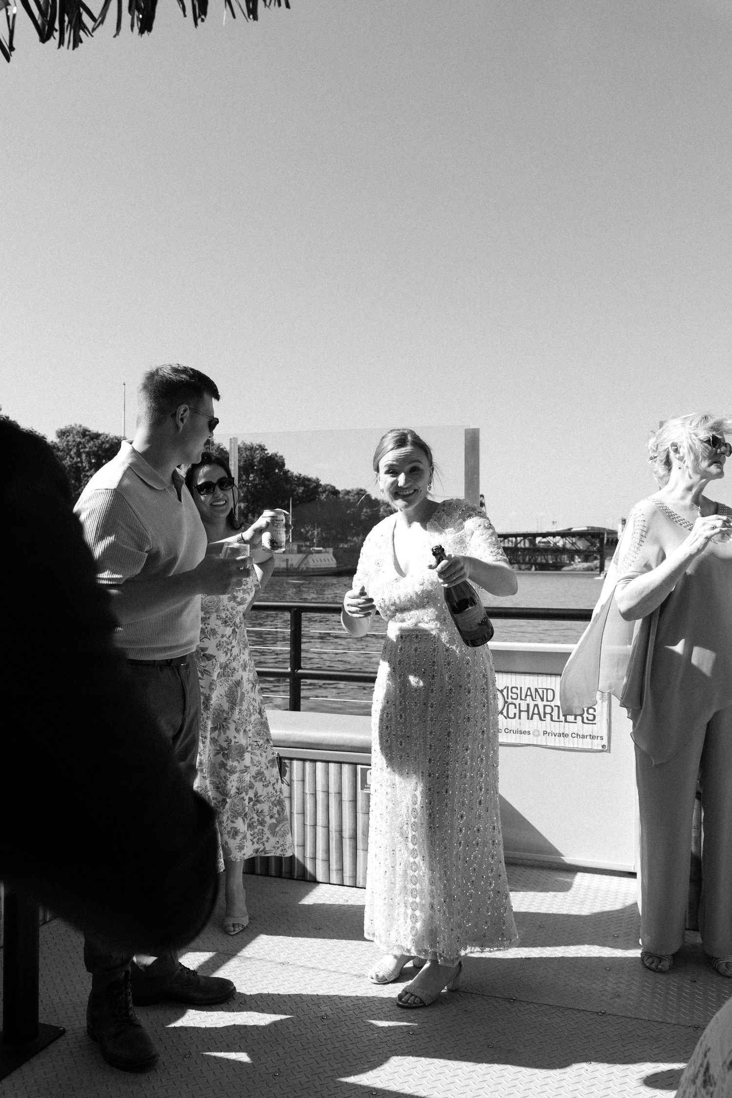 Black and white photo of guests celebrating outdoors near a waterway, woman in white lace dress holding champagne bottle.