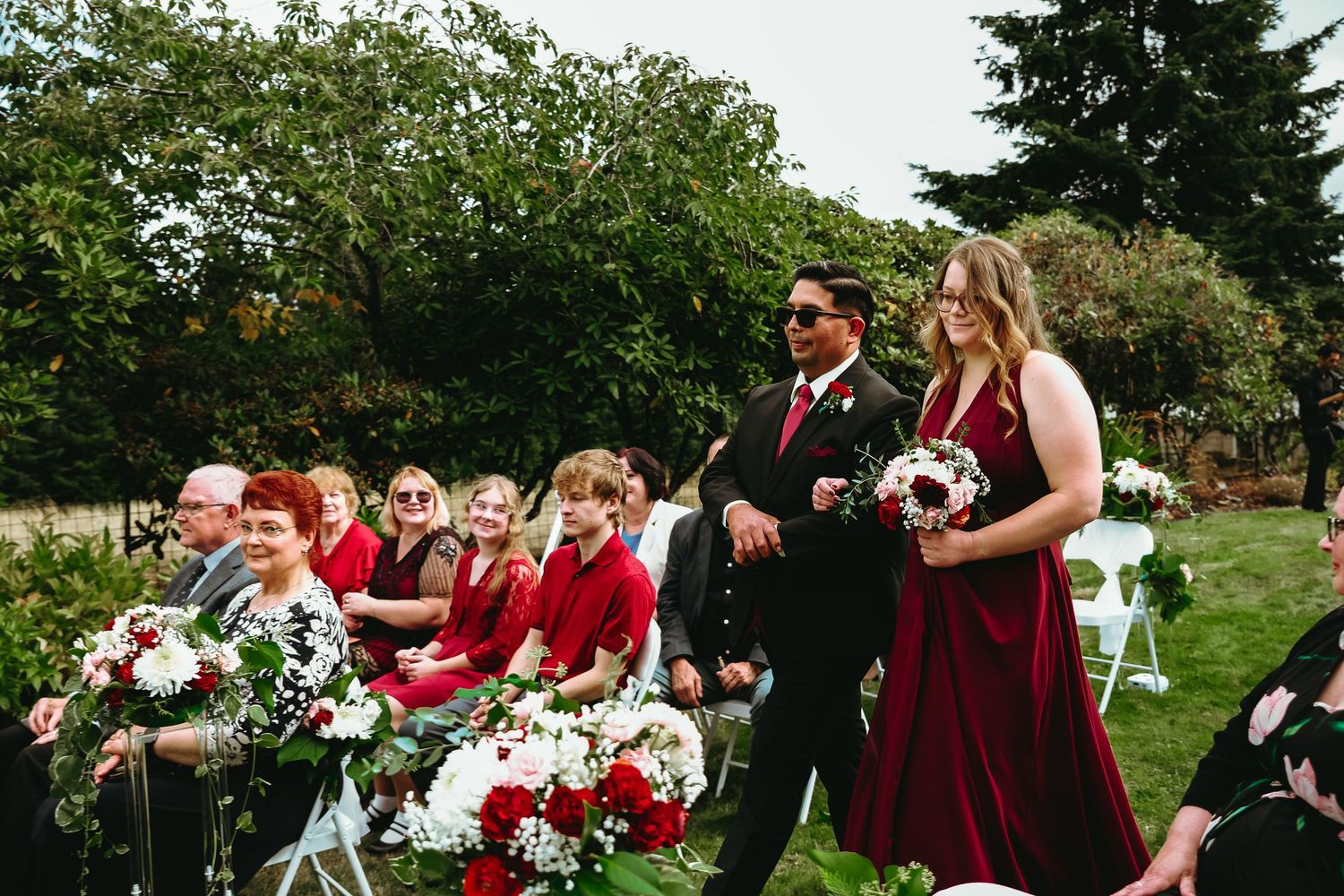 Groom in a dark suit and sunglasses walks a bridesmaid in a burgundy gown holding a bouquet past guests seated at an outdoor garden wedding.
