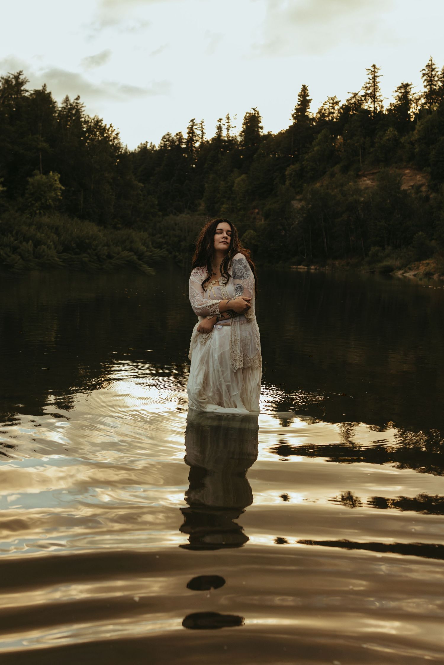 A woman in a white dress stands waist-deep in a calm lake surrounded by dark evergreen trees at dusk.