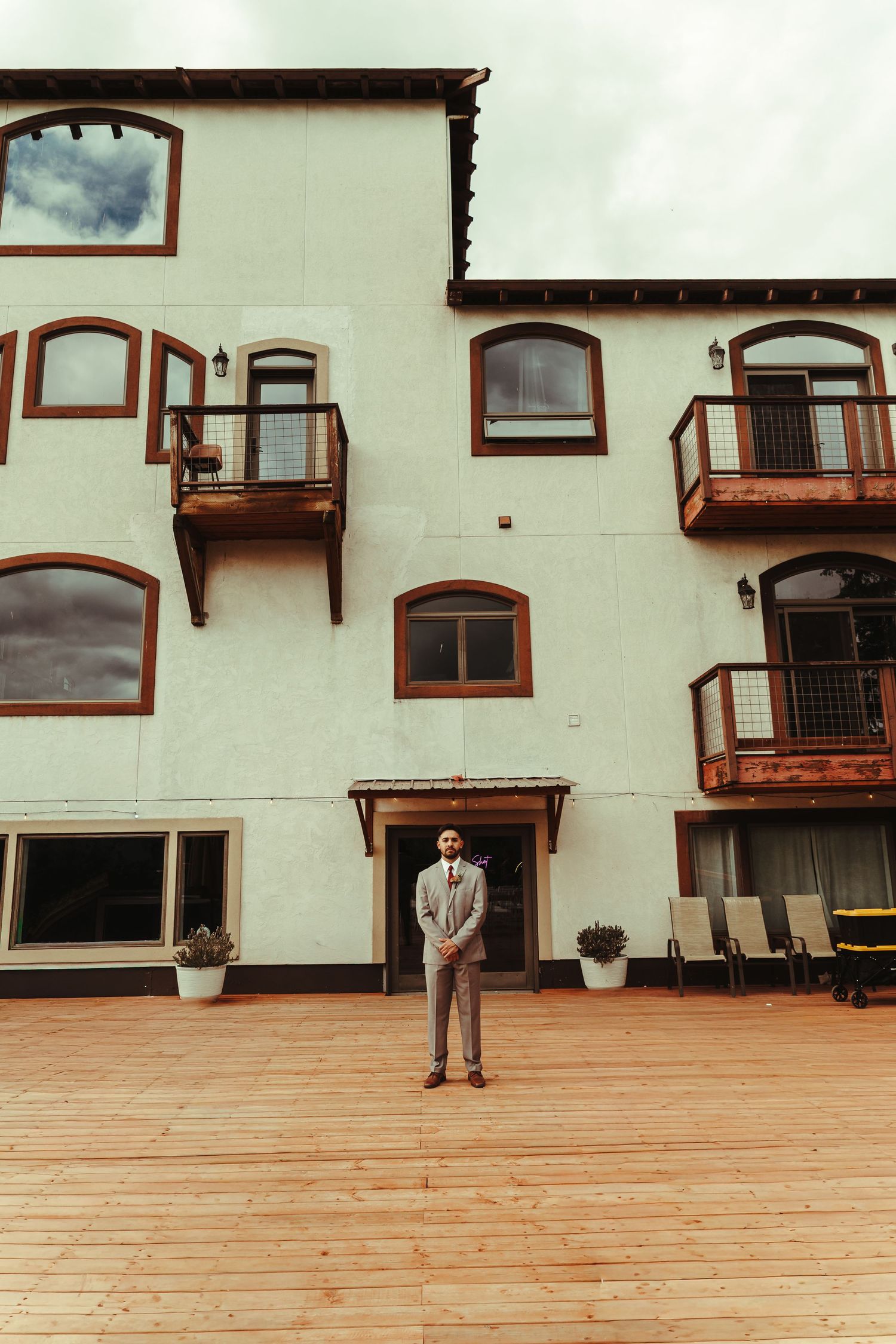 Groom in gray suit stands on wooden deck before elegant white building with balconies.