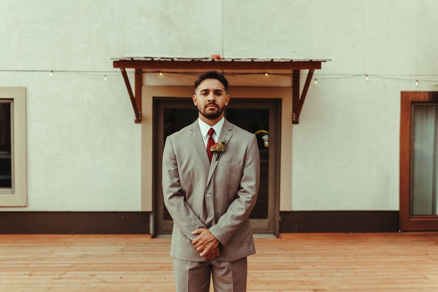 Groom in gray suit with floral boutonniere stands confidently outside a venue on a wooden deck.