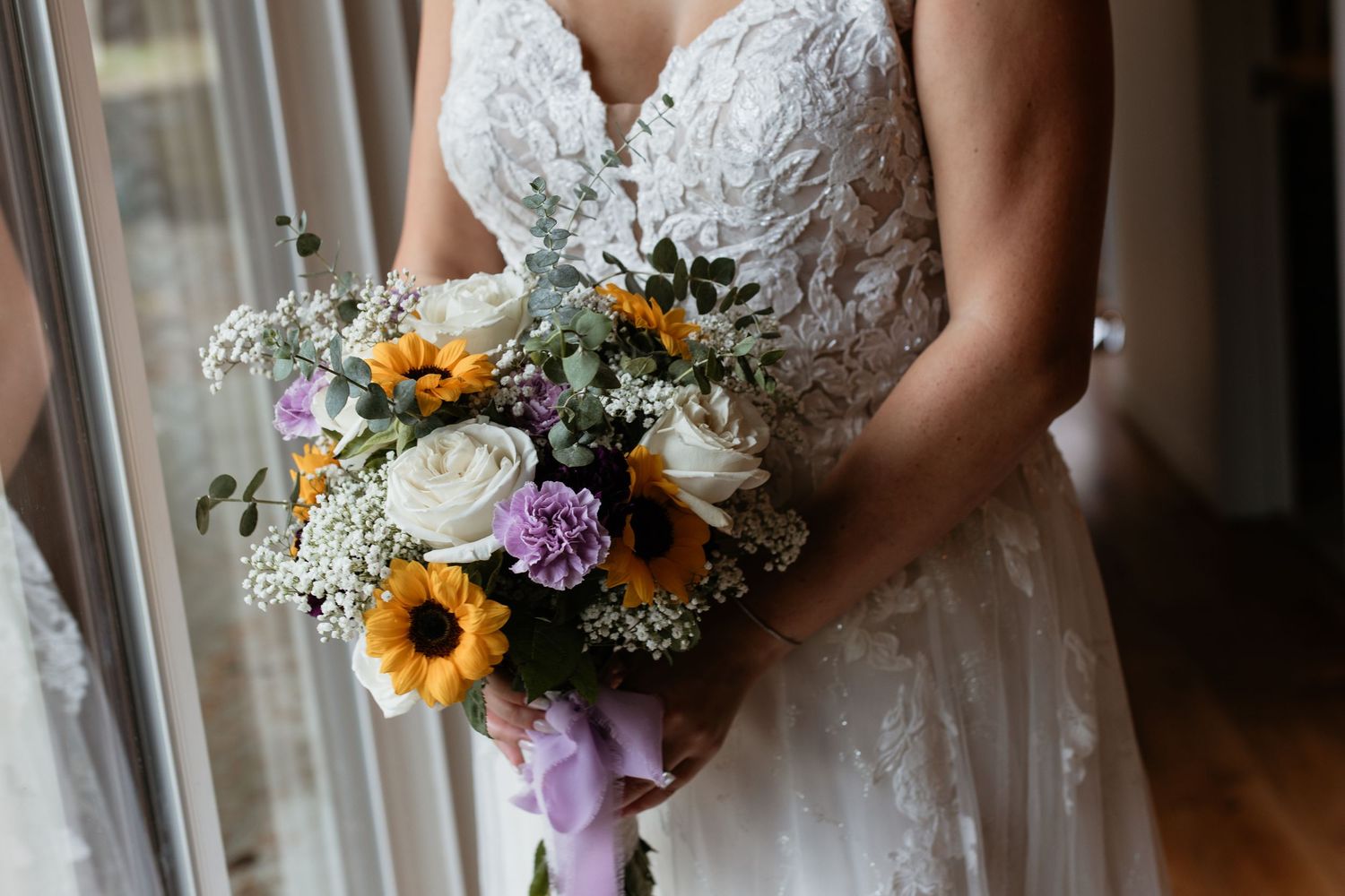 Bride holding colorful wedding bouquet with sunflowers, white roses, and purple blooms.