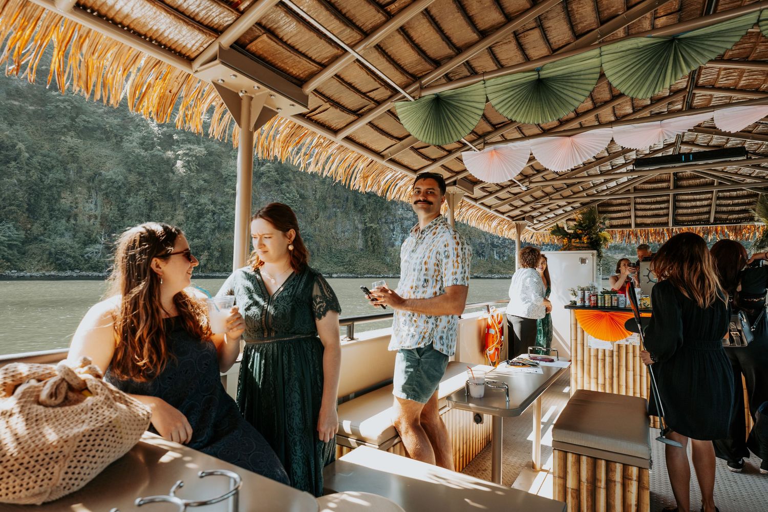 People socializing on a tropical tiki boat with bamboo decor, thatched roof, and scenic river views.