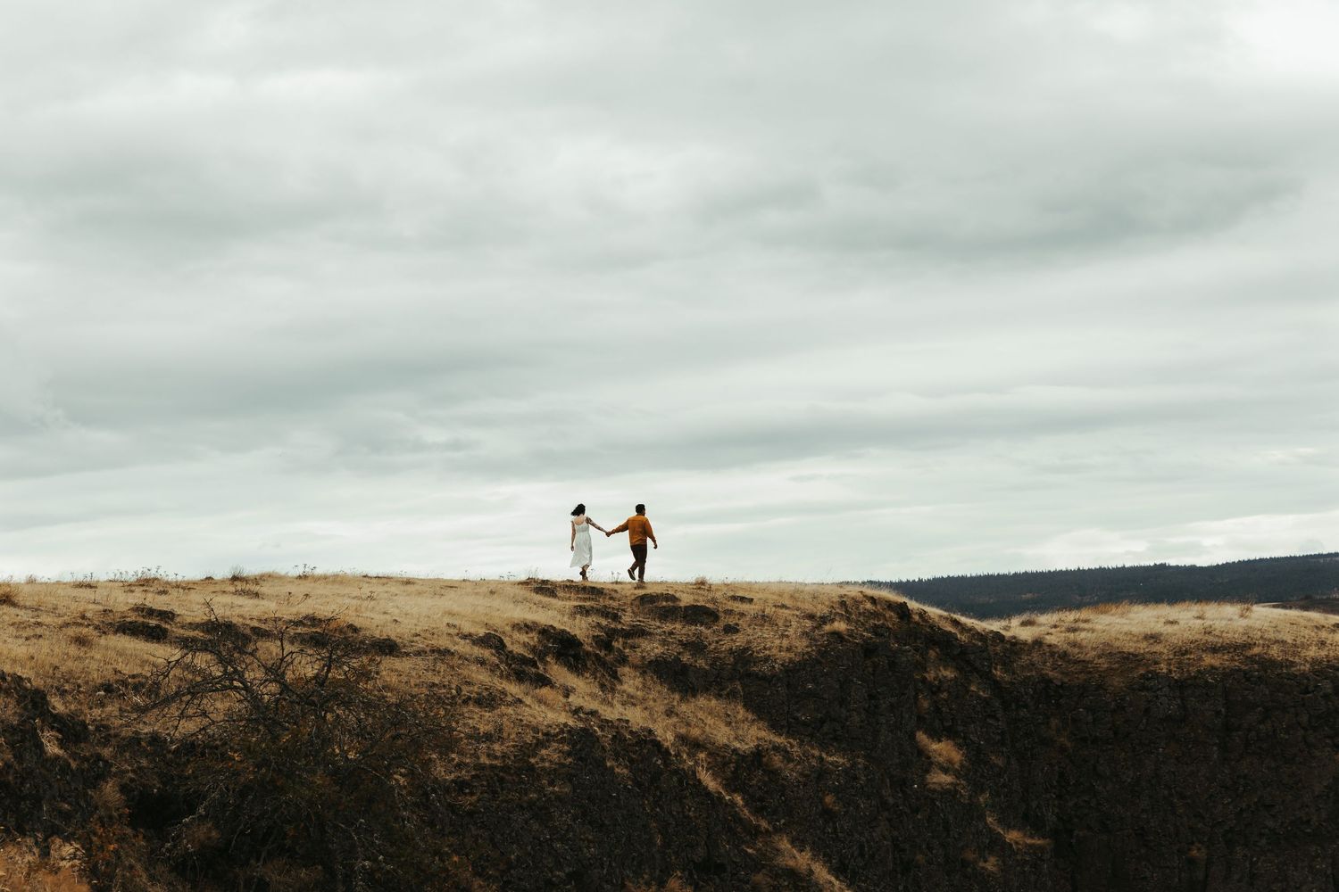 Couple holding hands on a rugged hilltop under a dramatic cloudy sky in an open landscape.