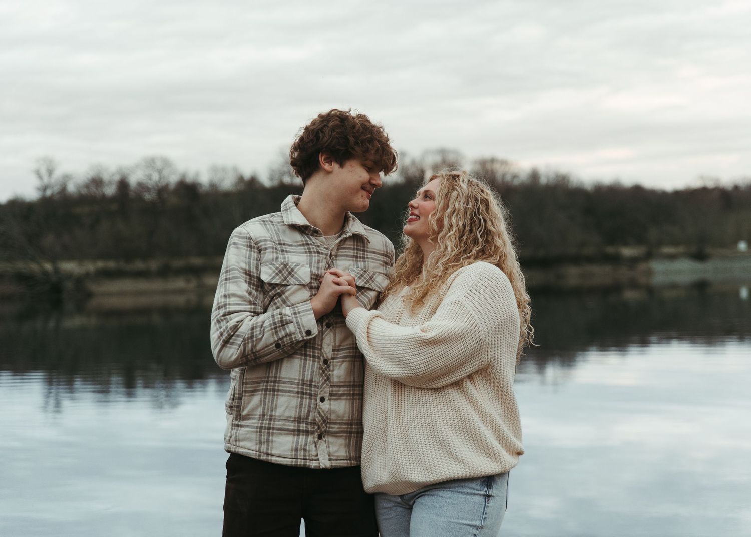 Young couple holding hands and smiling at each other by a reflective lake with trees in the background.