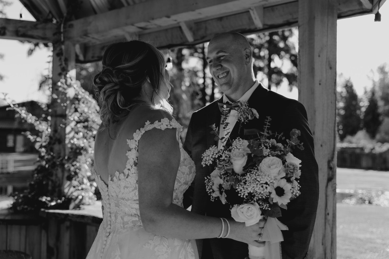 Black and white photo of groom smiling at bride holding floral bouquet during wedding first look.