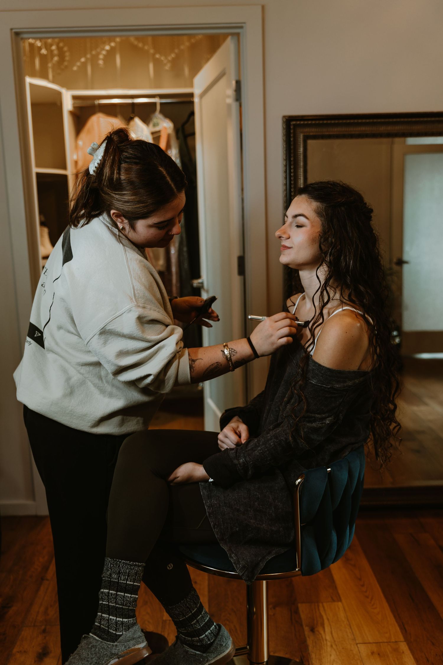 Bride having hairspray applied to her curly hair while seated before a mirror during bridal prep.