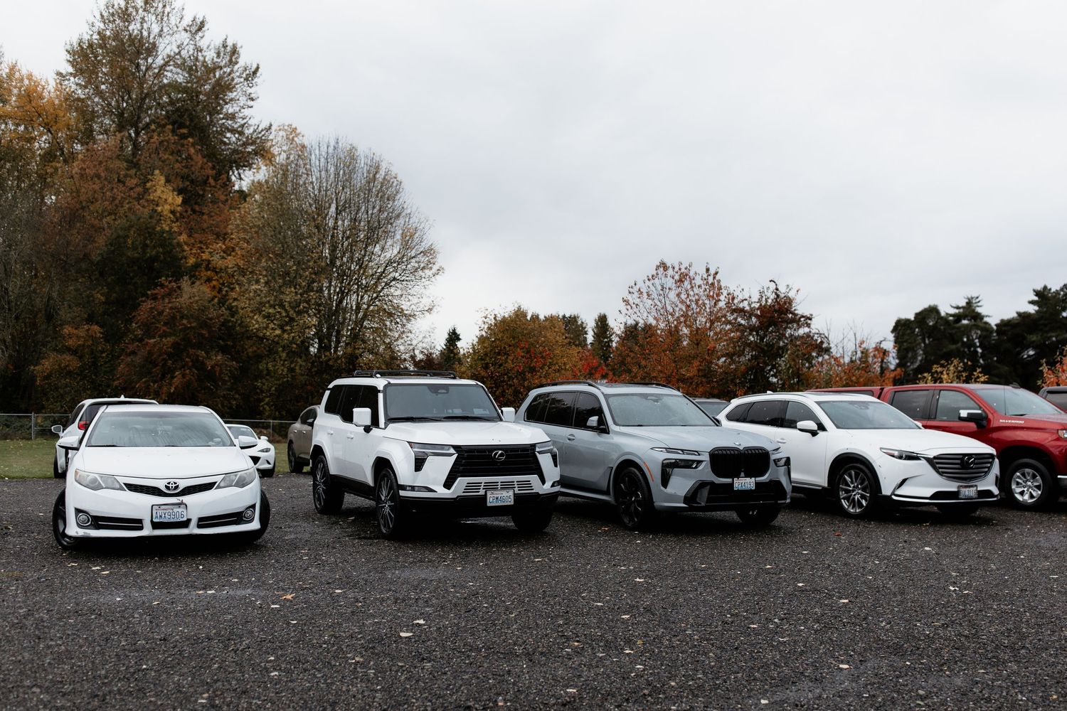 Row of SUVs and sedans parked in gravel lot surrounded by autumn foliage trees.