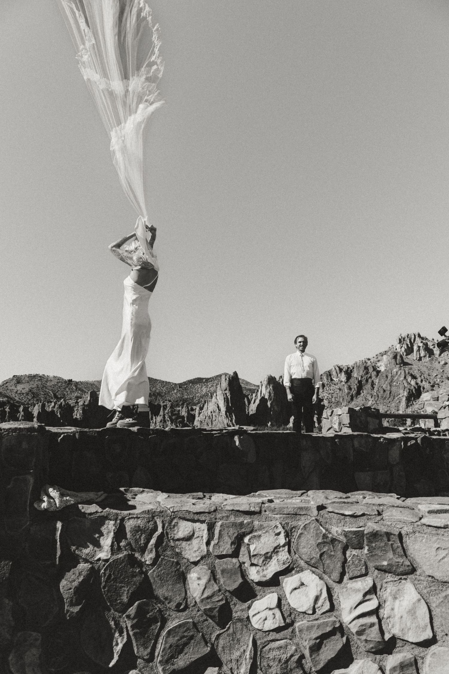 Black and white photo of a bride releasing a veil on stone ruins with groom standing nearby in desert landscape.