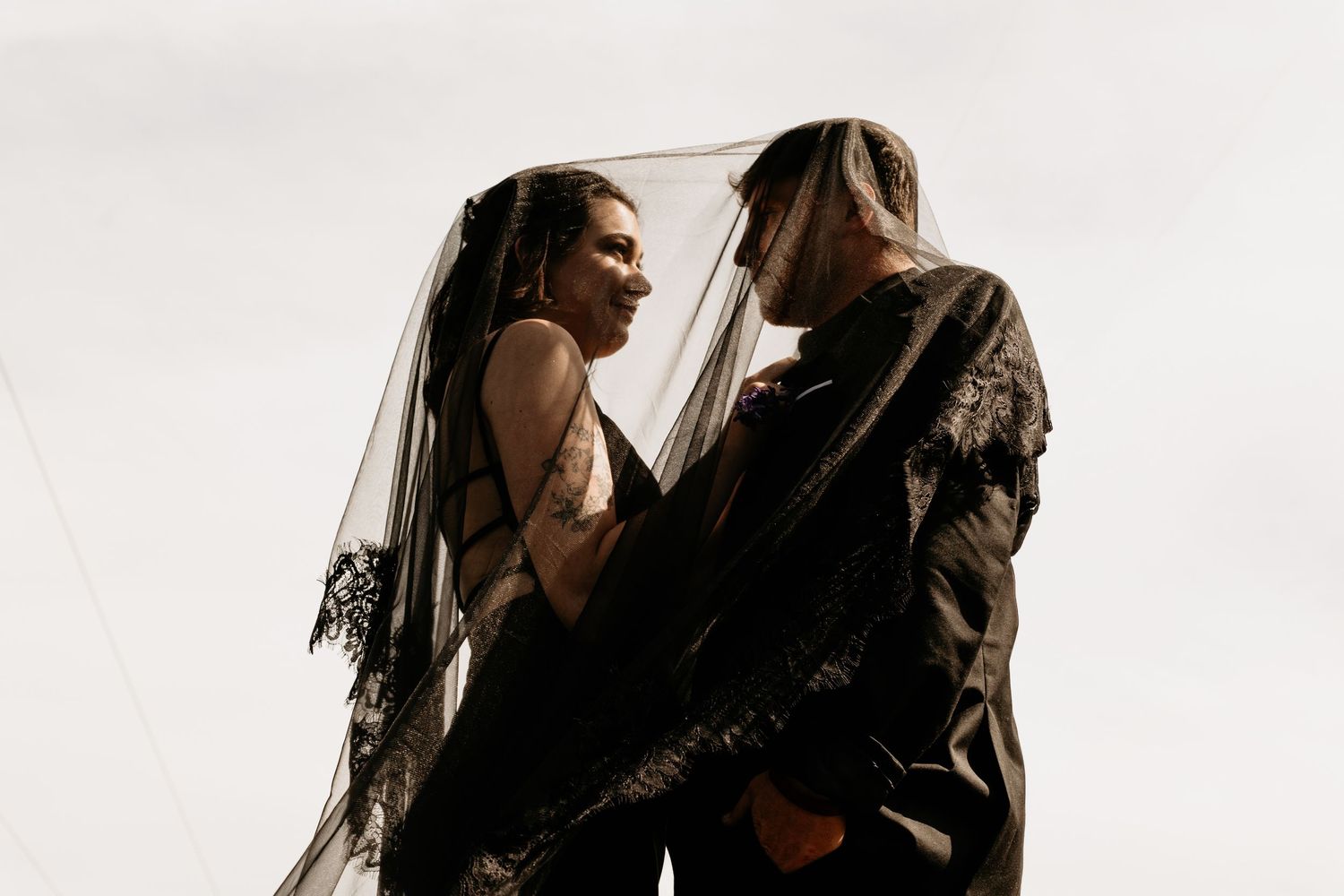 A couple shares an intimate moment under a black lace veil against a grey sky, dressed in elegant dark wedding attire.