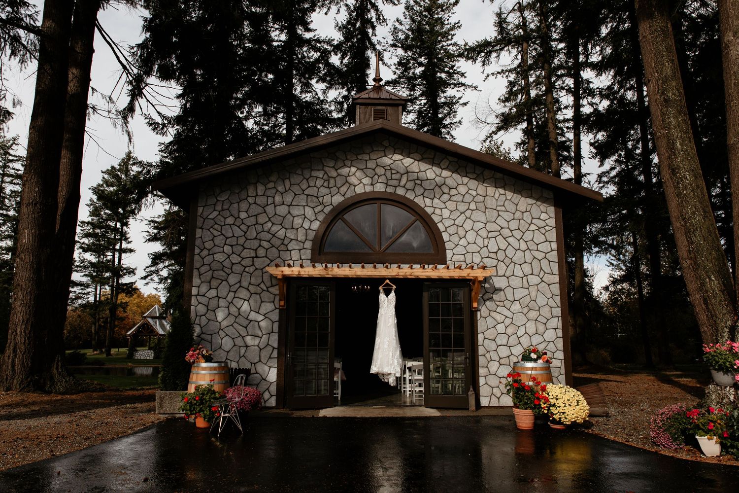 A wedding dress hangs in the doorway of a rustic stone chapel surrounded by tall evergreen trees.