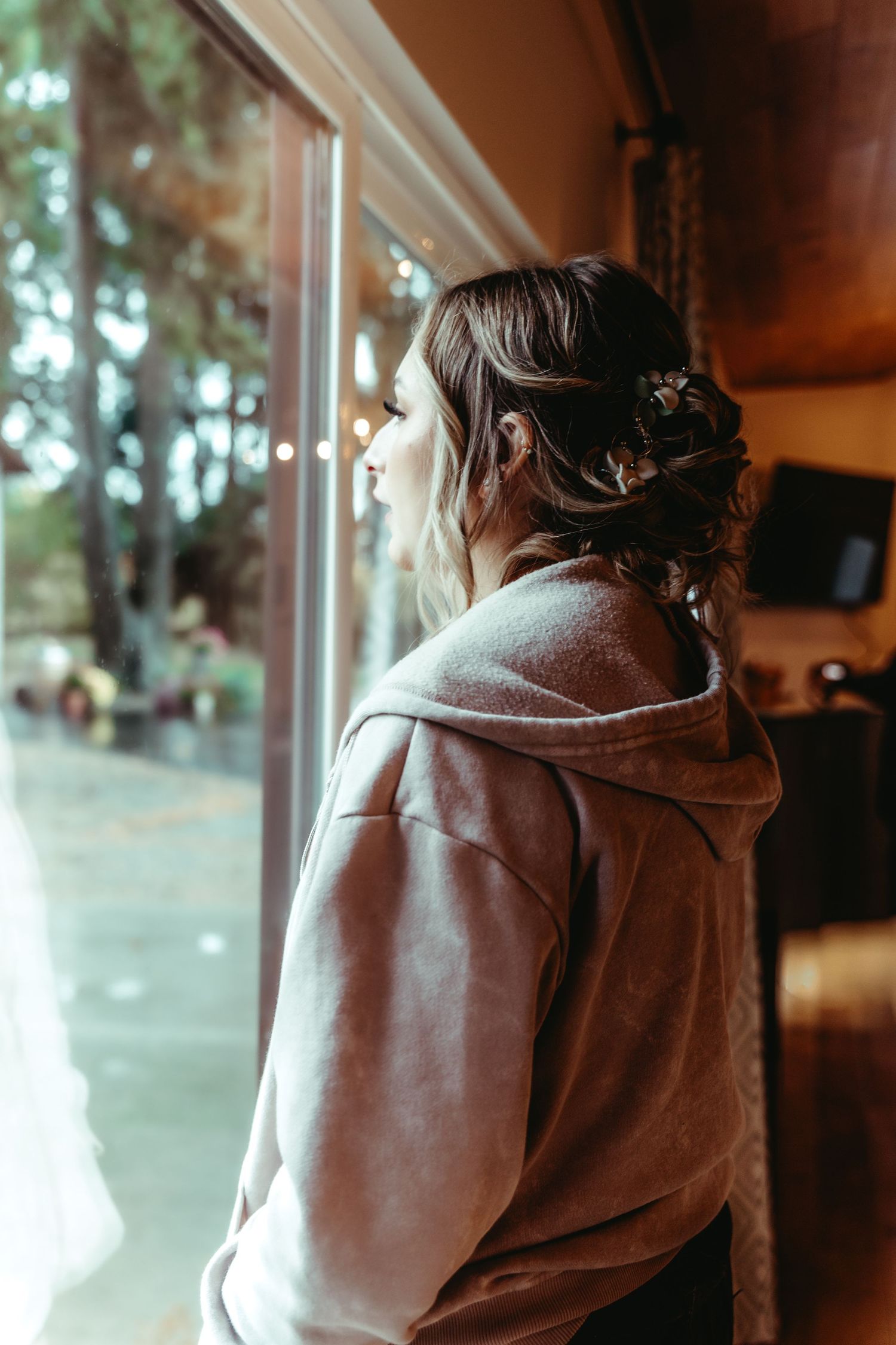 A woman in a gray hoodie with an elegant updo hairstyle gazes thoughtfully out a large window.