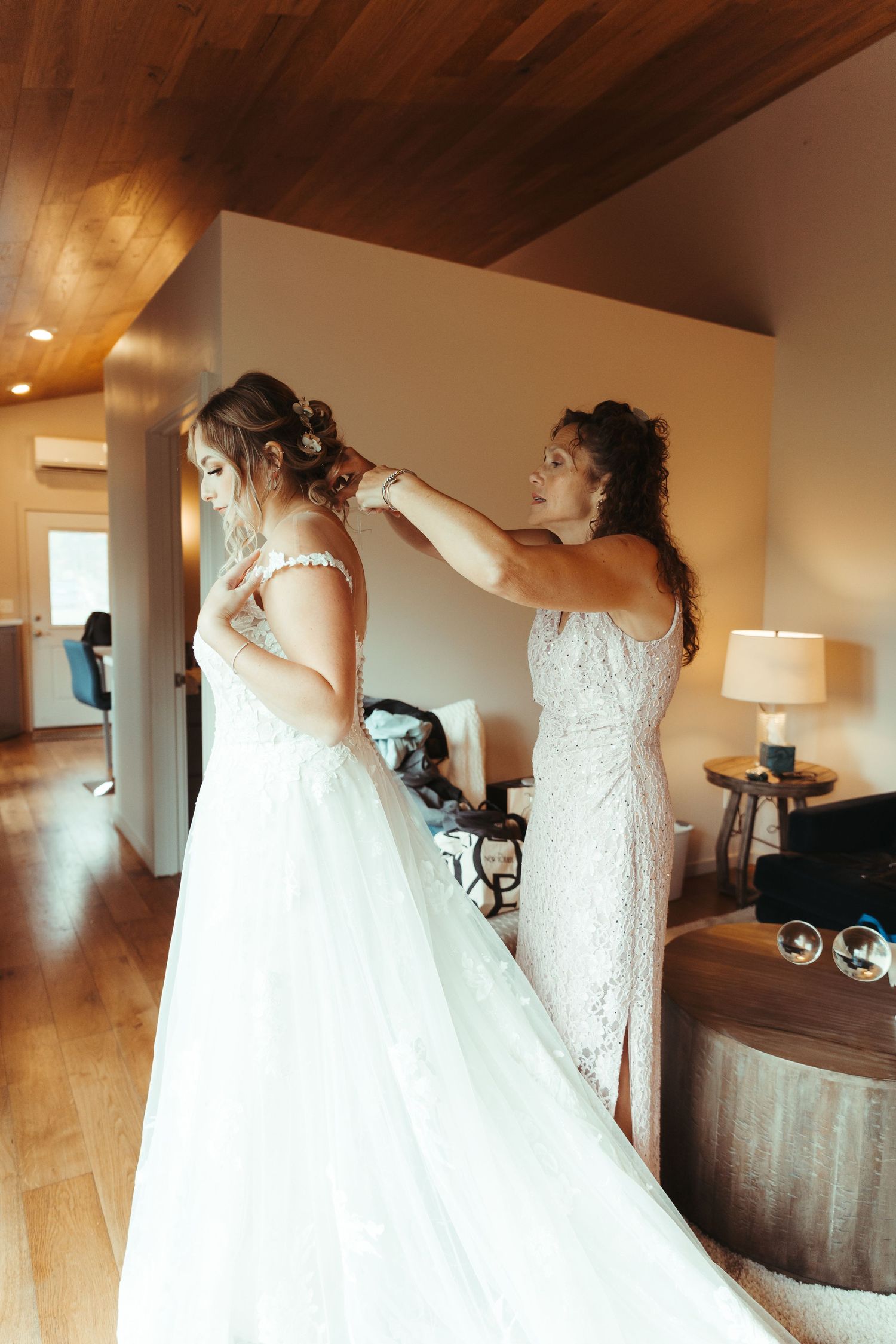 Curly-haired woman helping bride with back of elegant white lace wedding dress indoors.