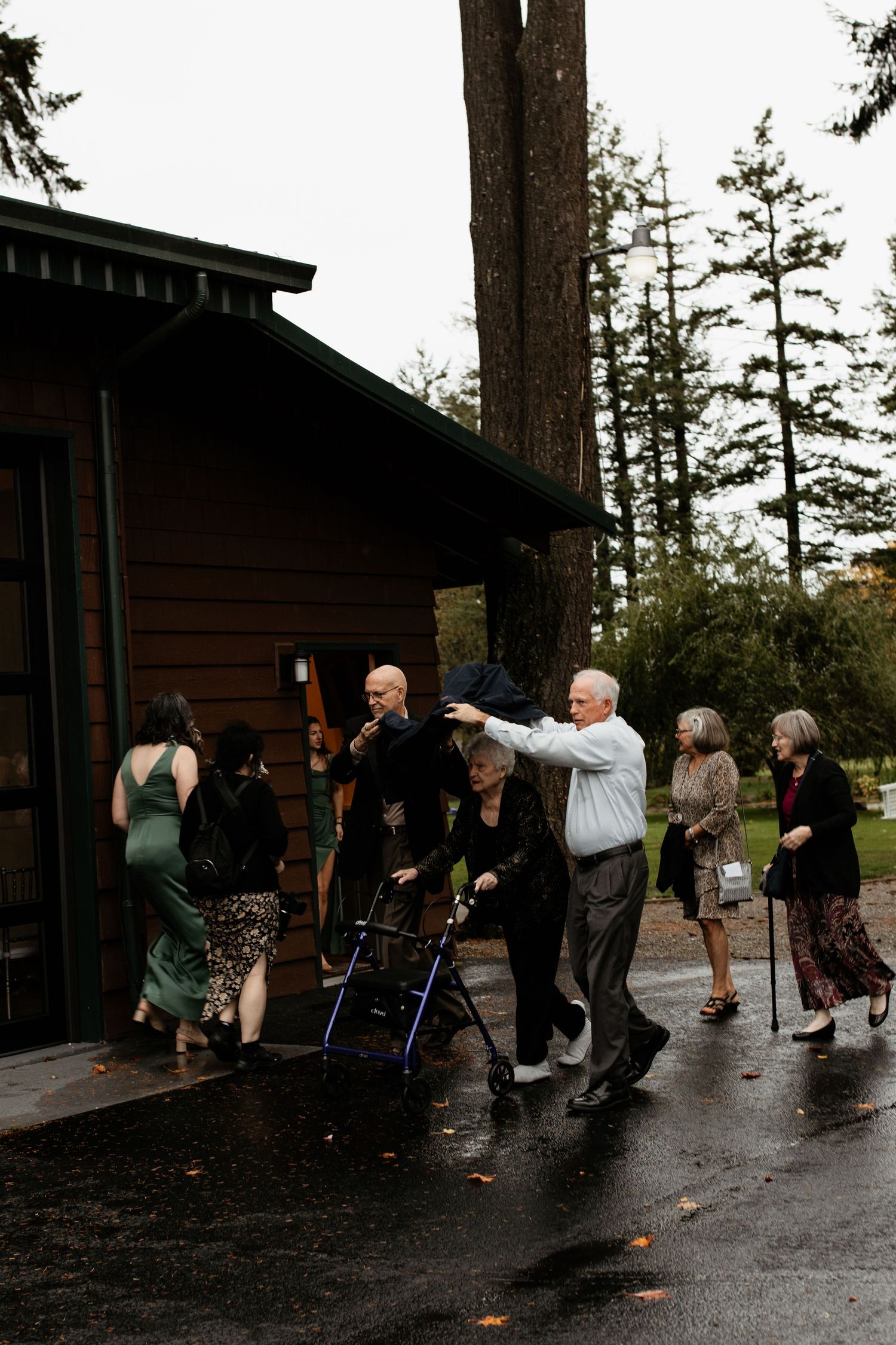 Wedding guests arriving in rain at rustic wooden venue with umbrella assistance and mobility walker.