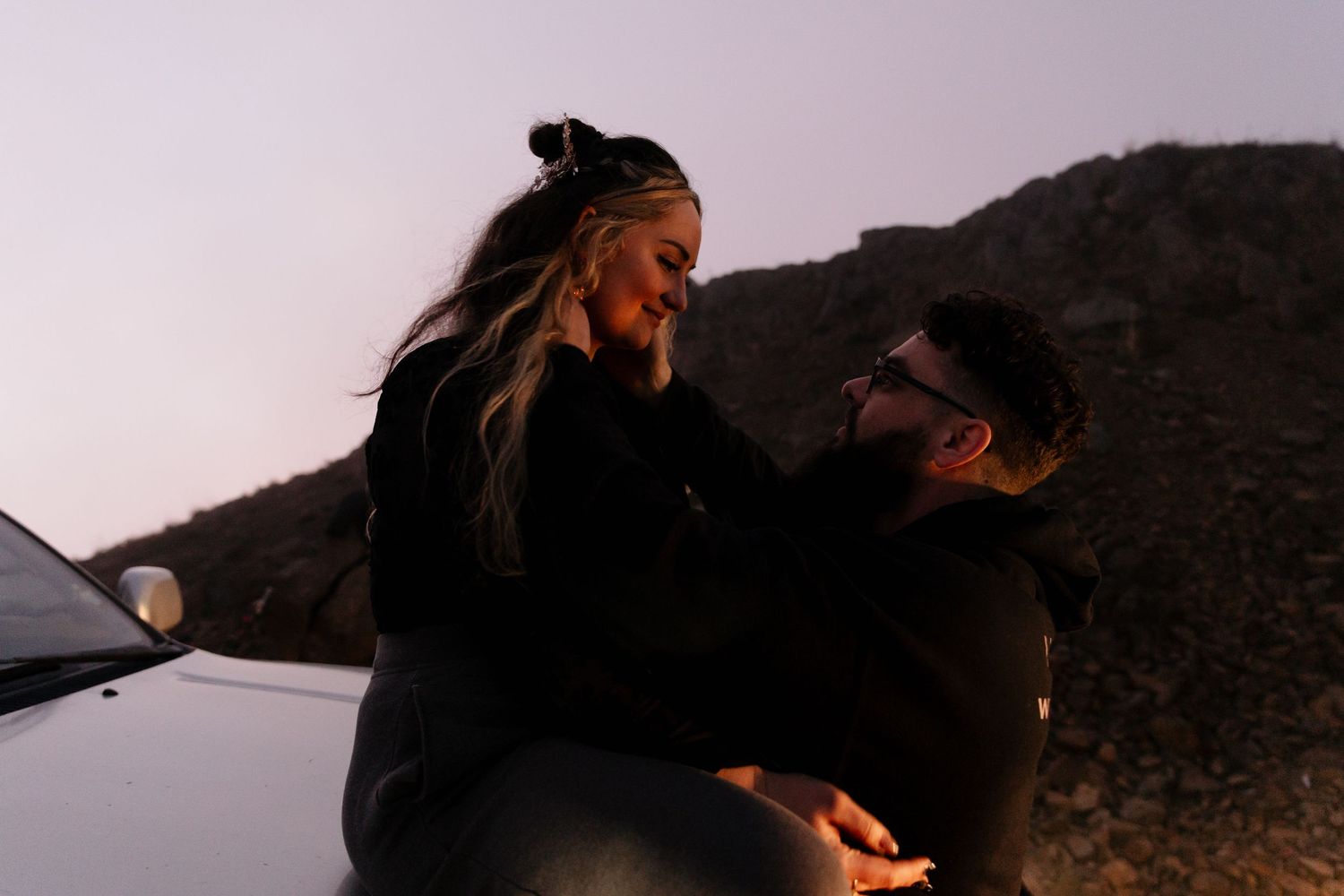 Romantic couple sharing an intimate moment sitting on a car hood at sunset in a rocky landscape.