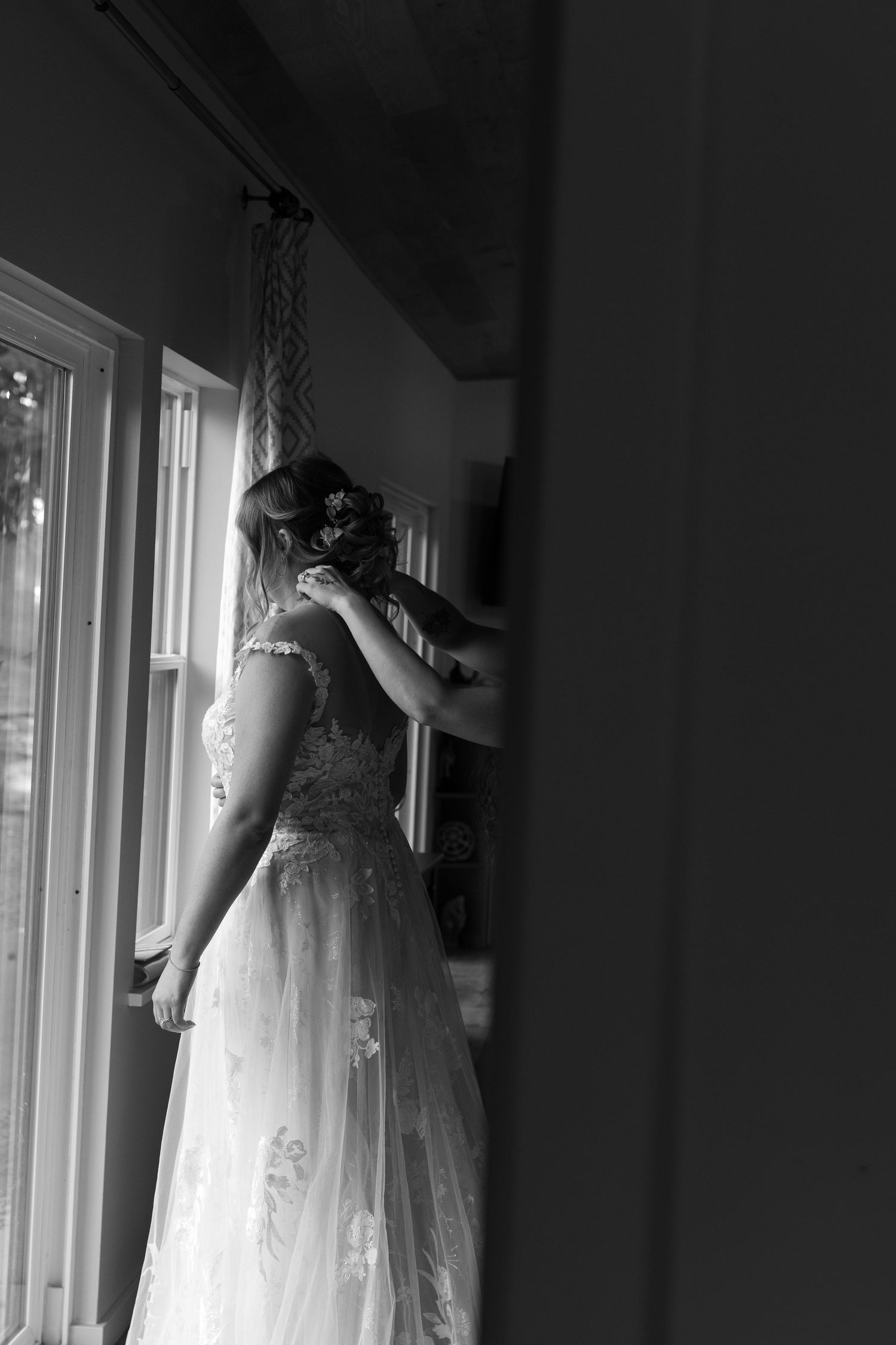 Black and white photo of bride in lace gown standing by window being helped by bridesmaid.