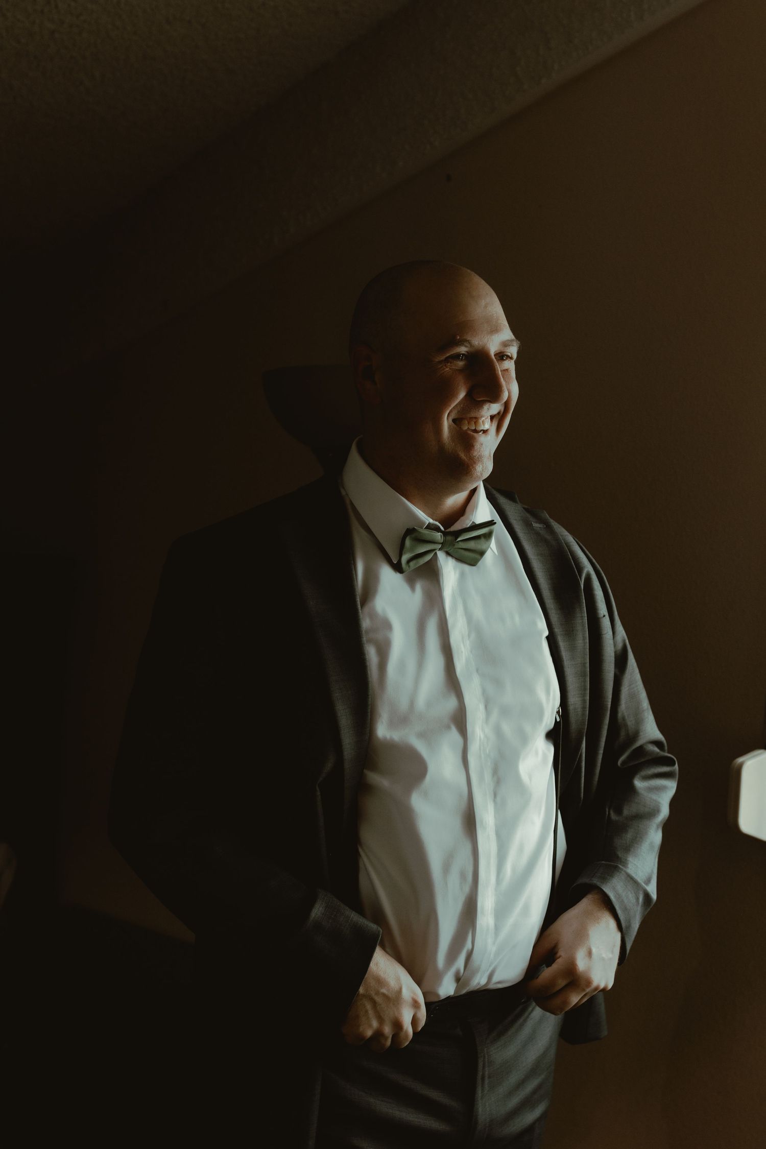 Smiling groom in dark suit and green bow tie adjusts suspenders in warm moody indoor lighting.
