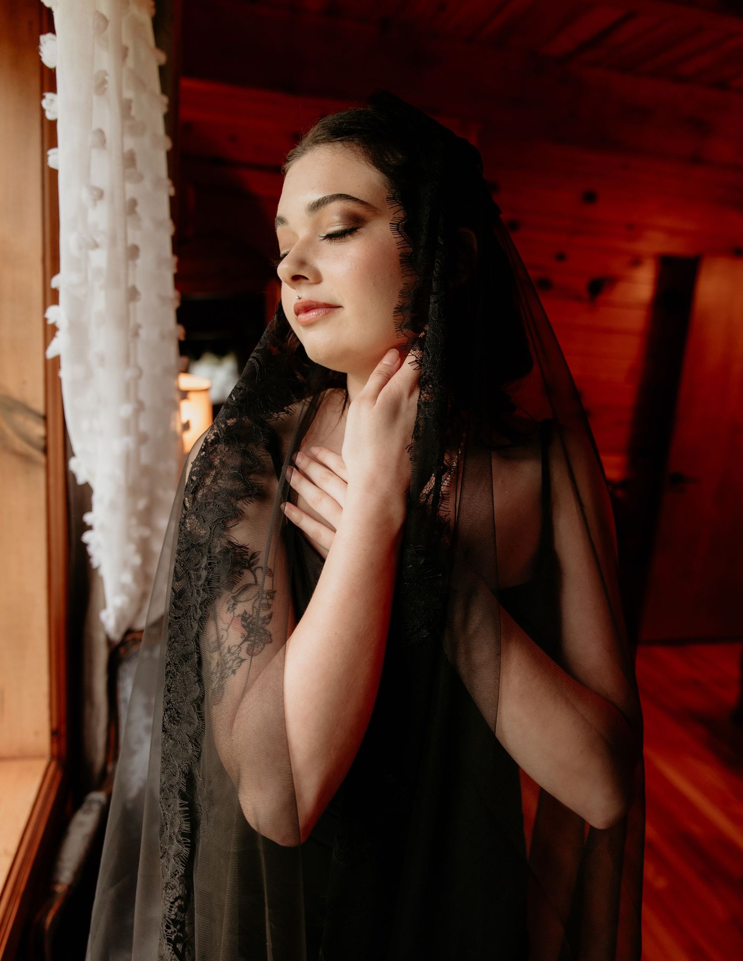 Woman in black lace veil poses gracefully by a window in a warm wooden interior setting.