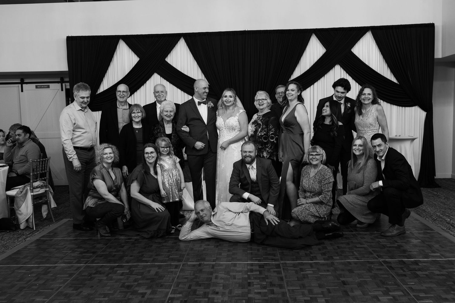 Black and white wedding group photo with bride and groom surrounded by family and friends before draped backdrop.