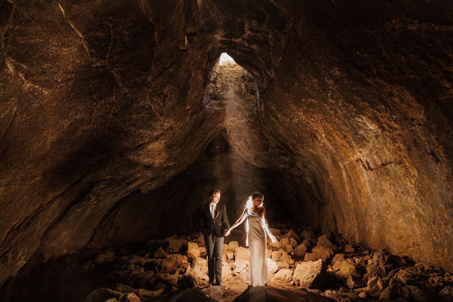 Couple holds hands in a dramatic cave wedding with light streaming through a rocky opening above them.
