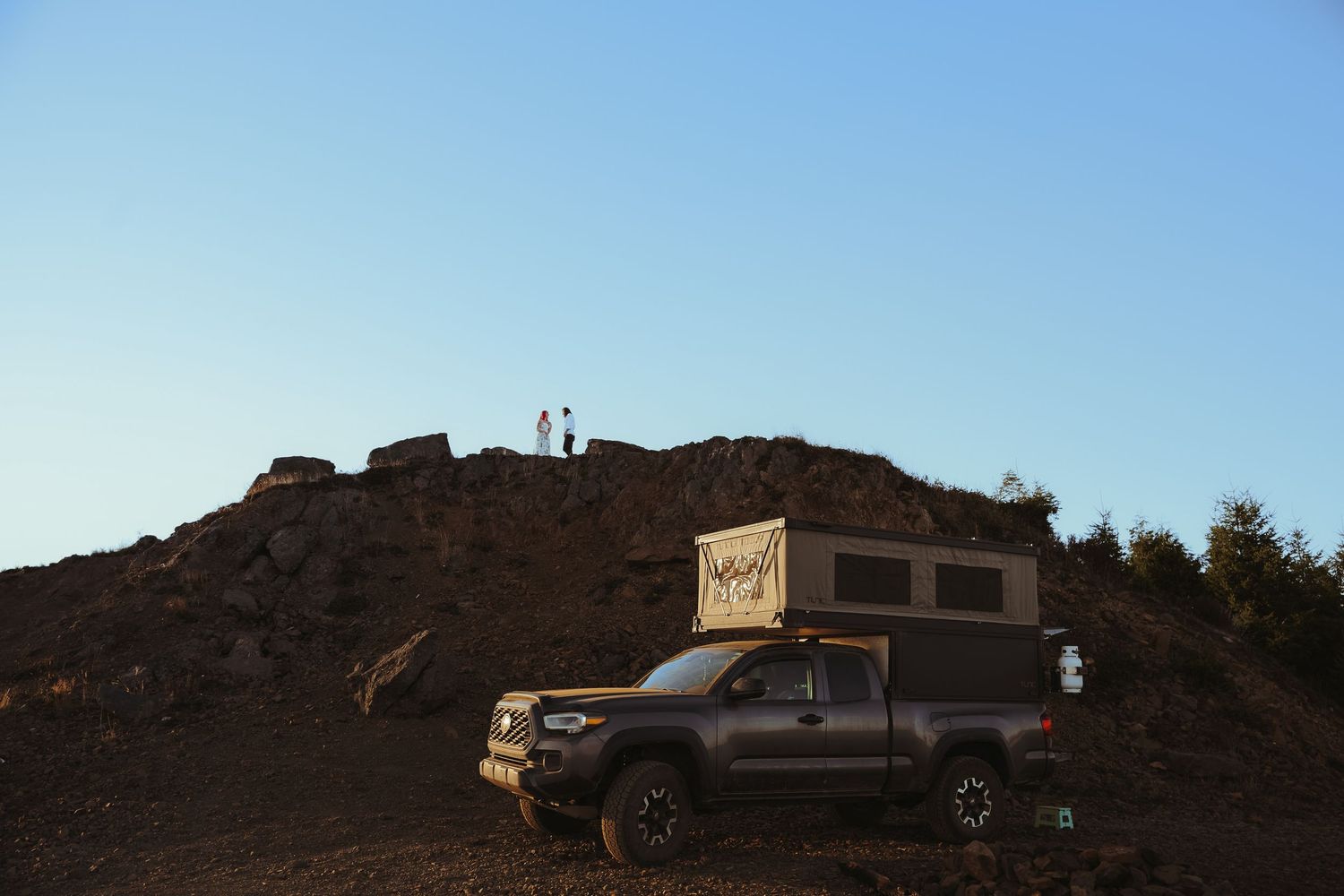 A gray Toyota Tacoma with a rooftop camper shell parked on a rocky volcanic landscape at dusk with two people on a hill.
