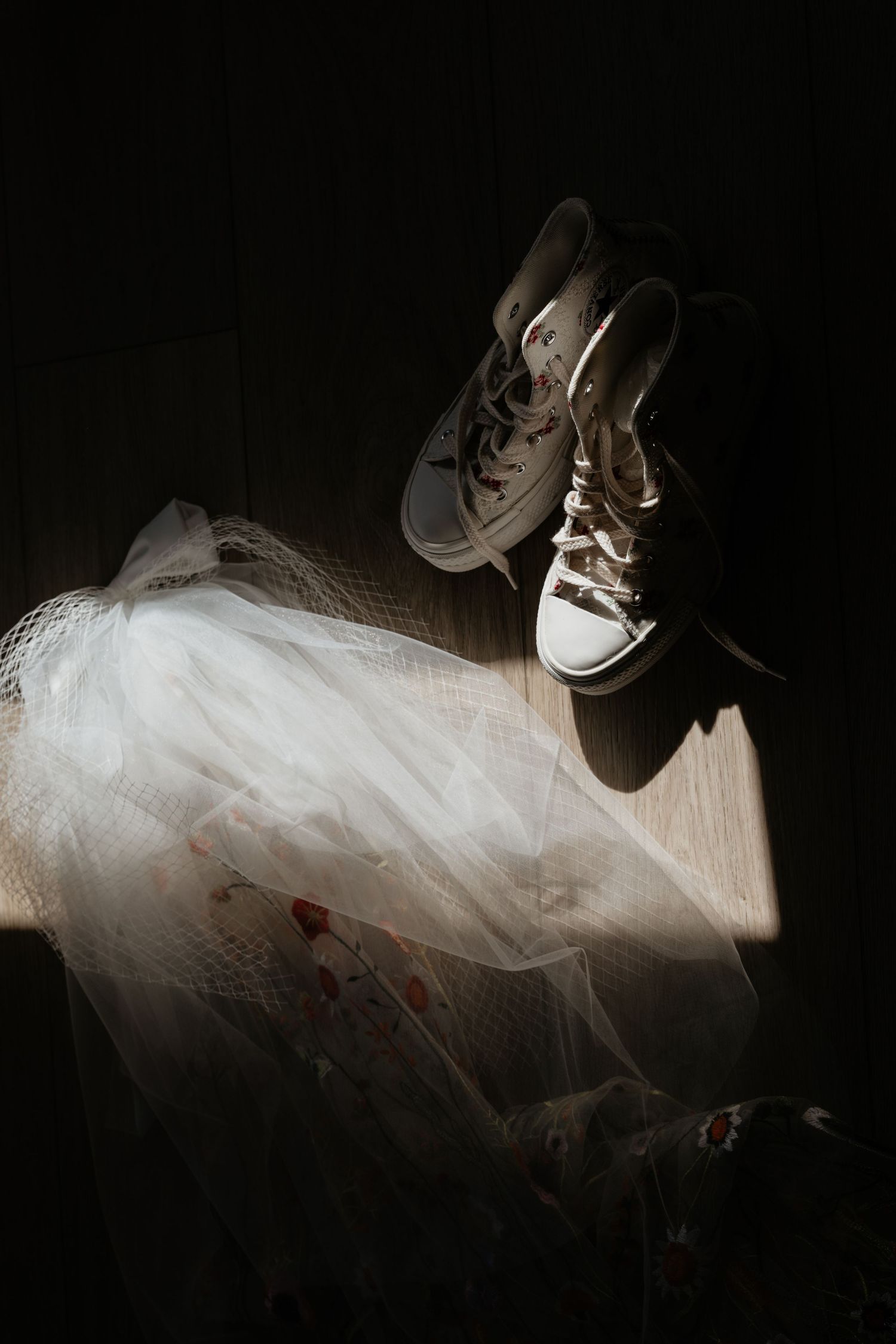 White Converse sneakers and a flowing tulle wedding dress illuminated by dramatic natural light on a wooden floor.